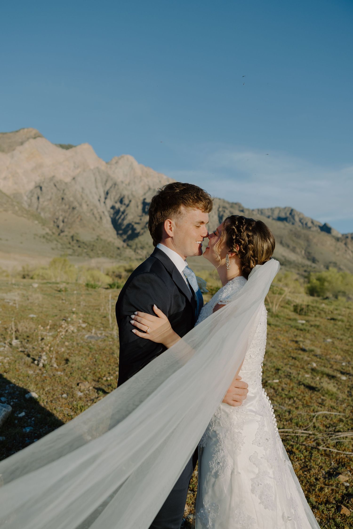 Bride and groom kissing with dramatic mountain backdrop and flowing veil on sunny wedding day.