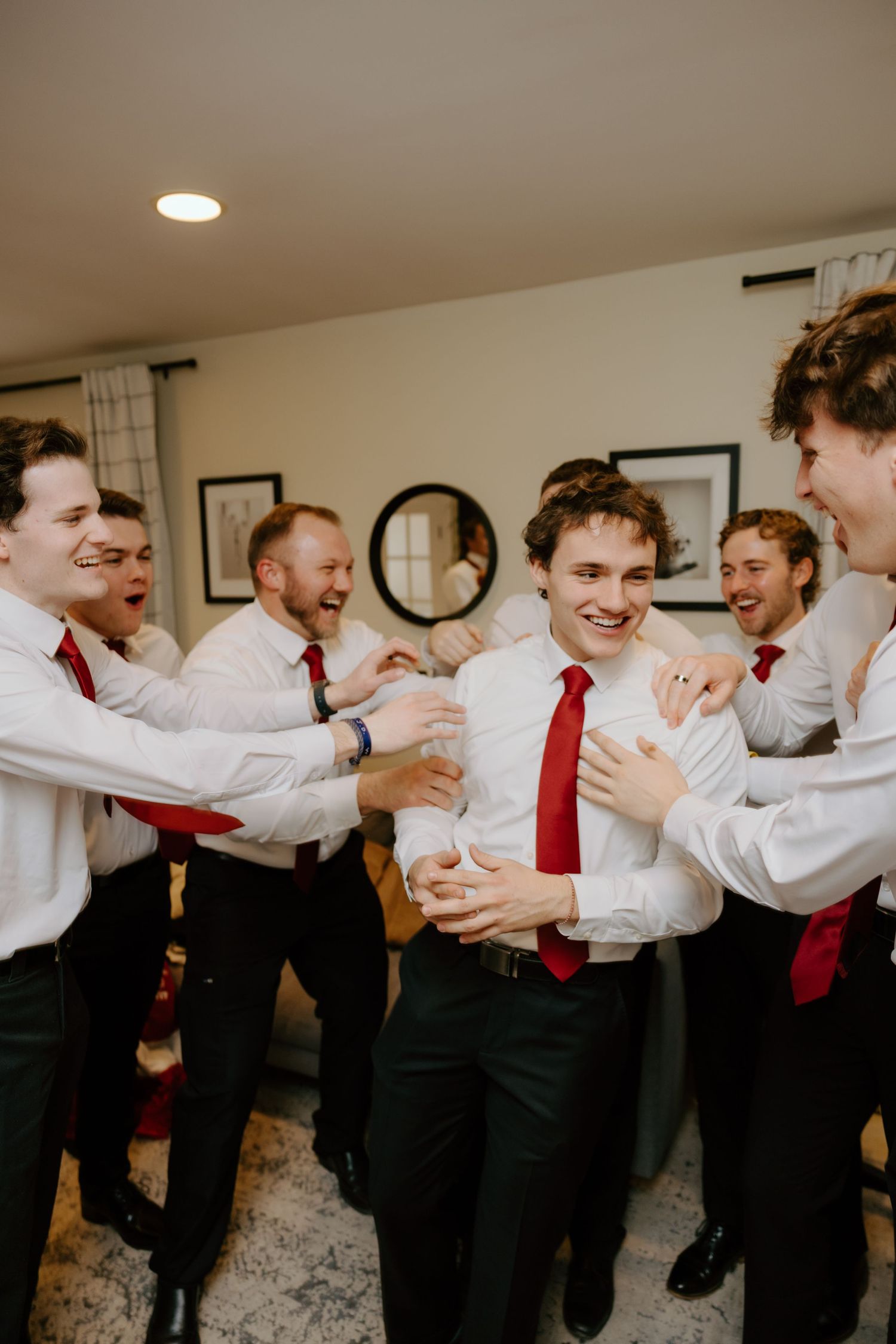 Excited groomsmen in white shirts and red ties celebrate with the groom during wedding day preparations indoors.