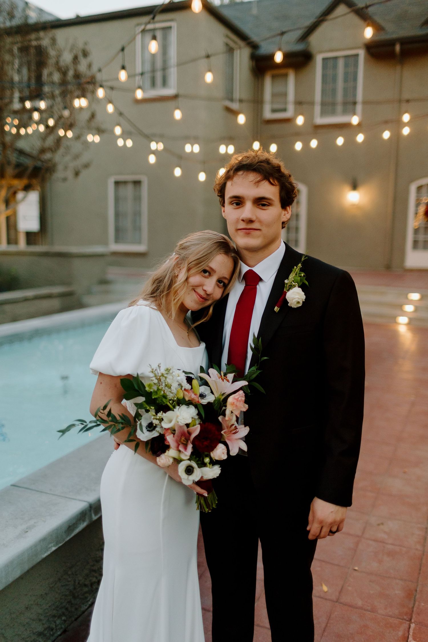 Bride in white gown holding pink and burgundy bouquet leans on groom in black suit with red tie near pool.