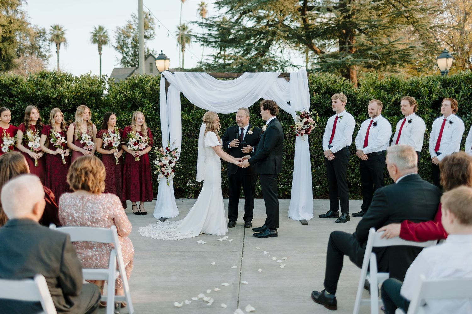 Outdoor wedding ceremony with bride and groom at floral arch, bridesmaids in burgundy dresses and groomsmen in suits.