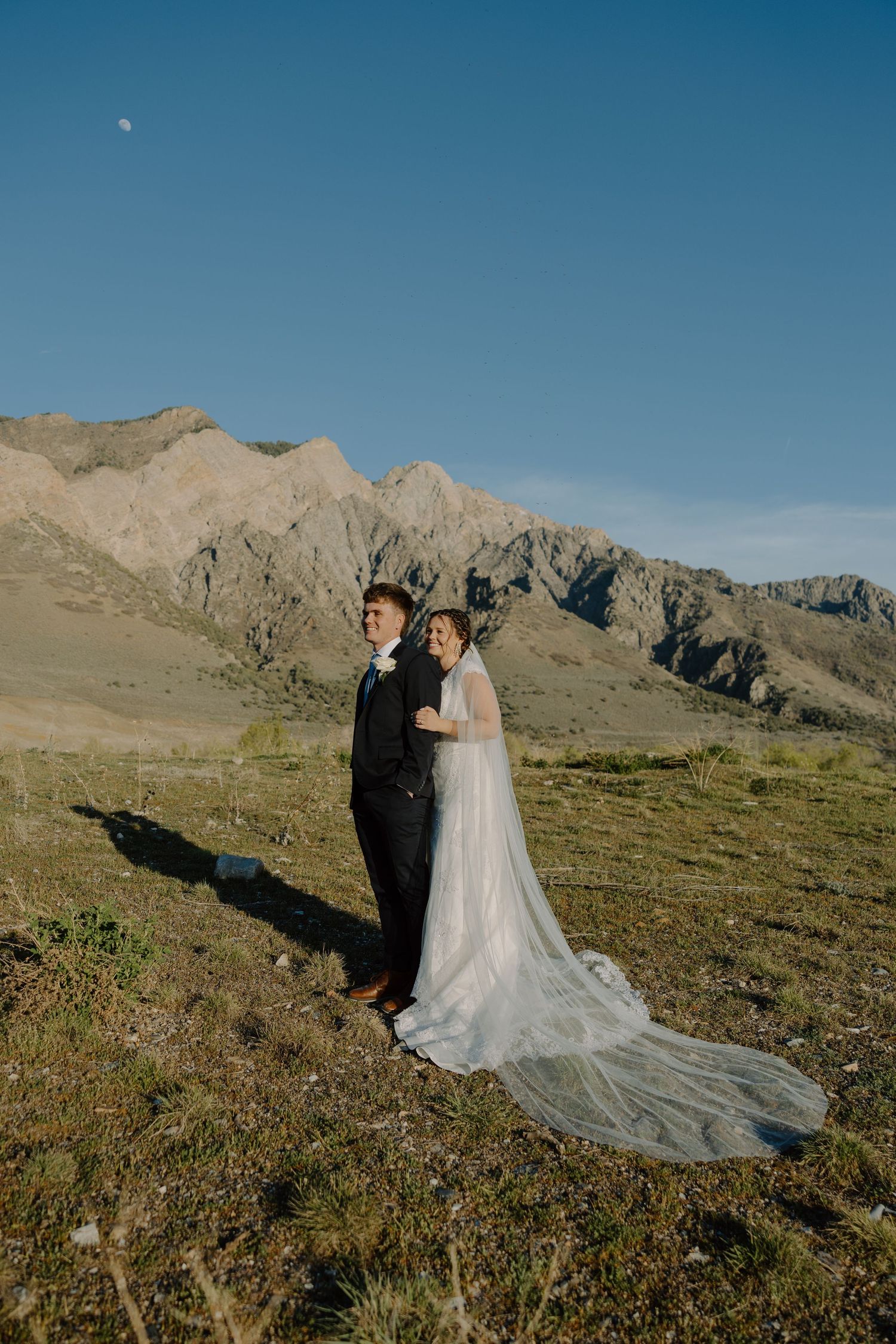 Bride in flowing white gown with train and groom stand together in a scenic mountain landscape.