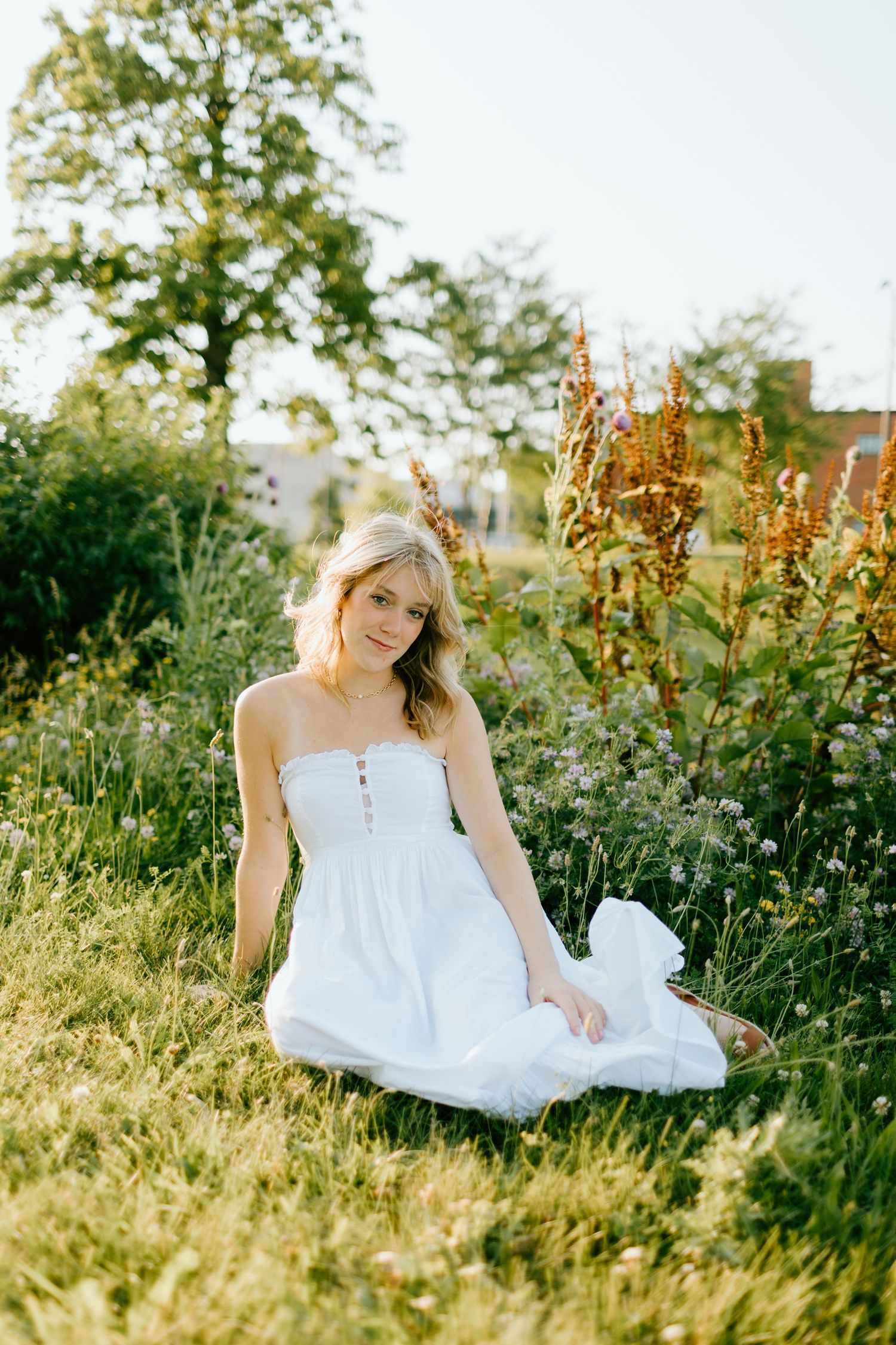 Young blonde woman in white strapless dress sitting gracefully in a sunlit garden surrounded by wildflowers and greenery.