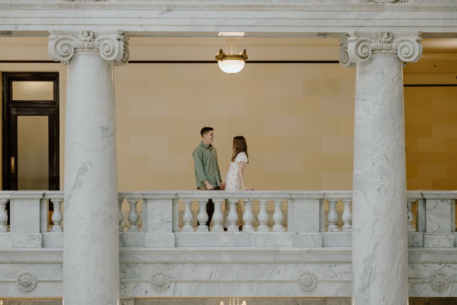 Couple standing together on a marble balcony between elegant white columns inside a grand classical building.