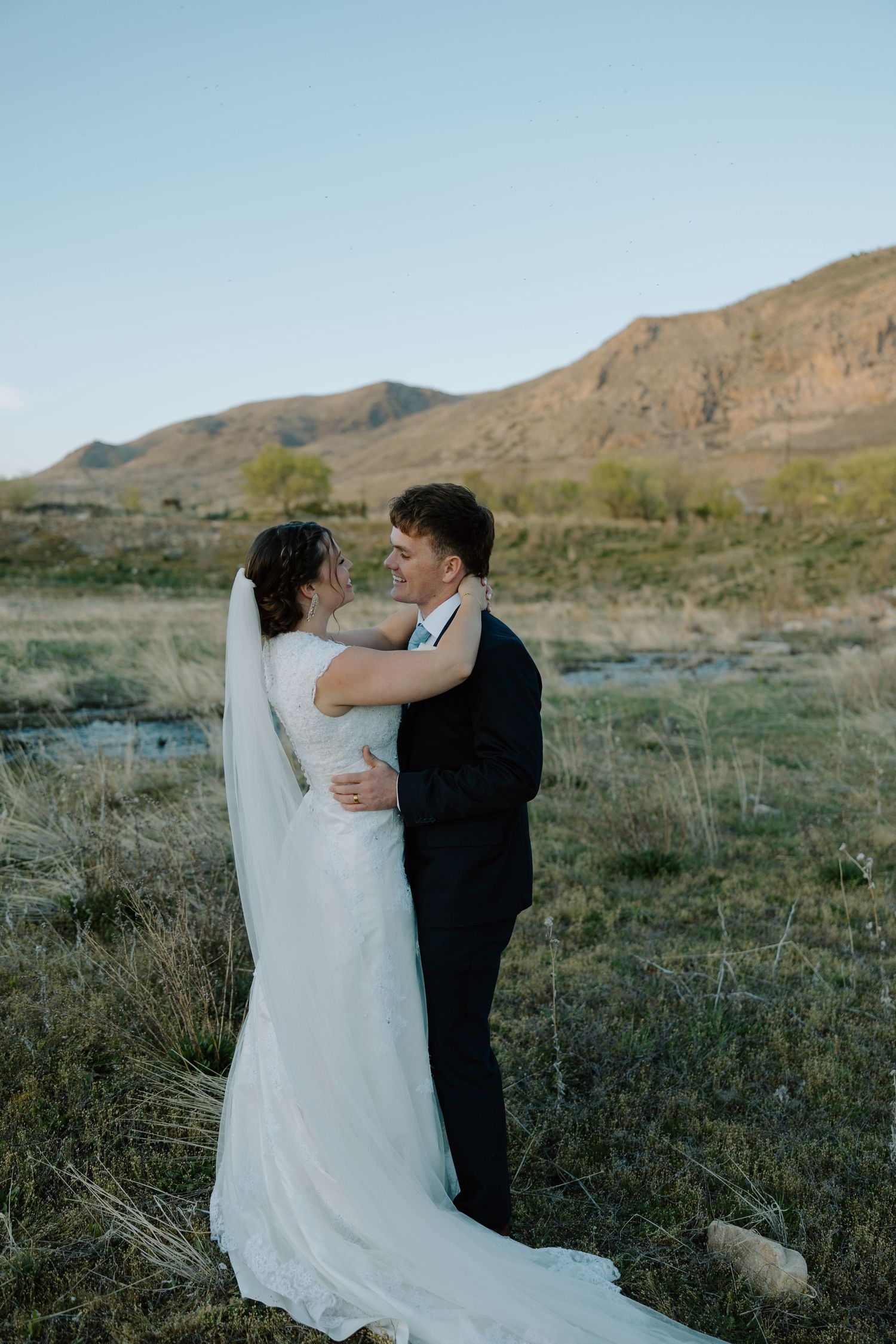 Bride and groom embrace in a golden field with mountain backdrop during outdoor wedding portrait session.