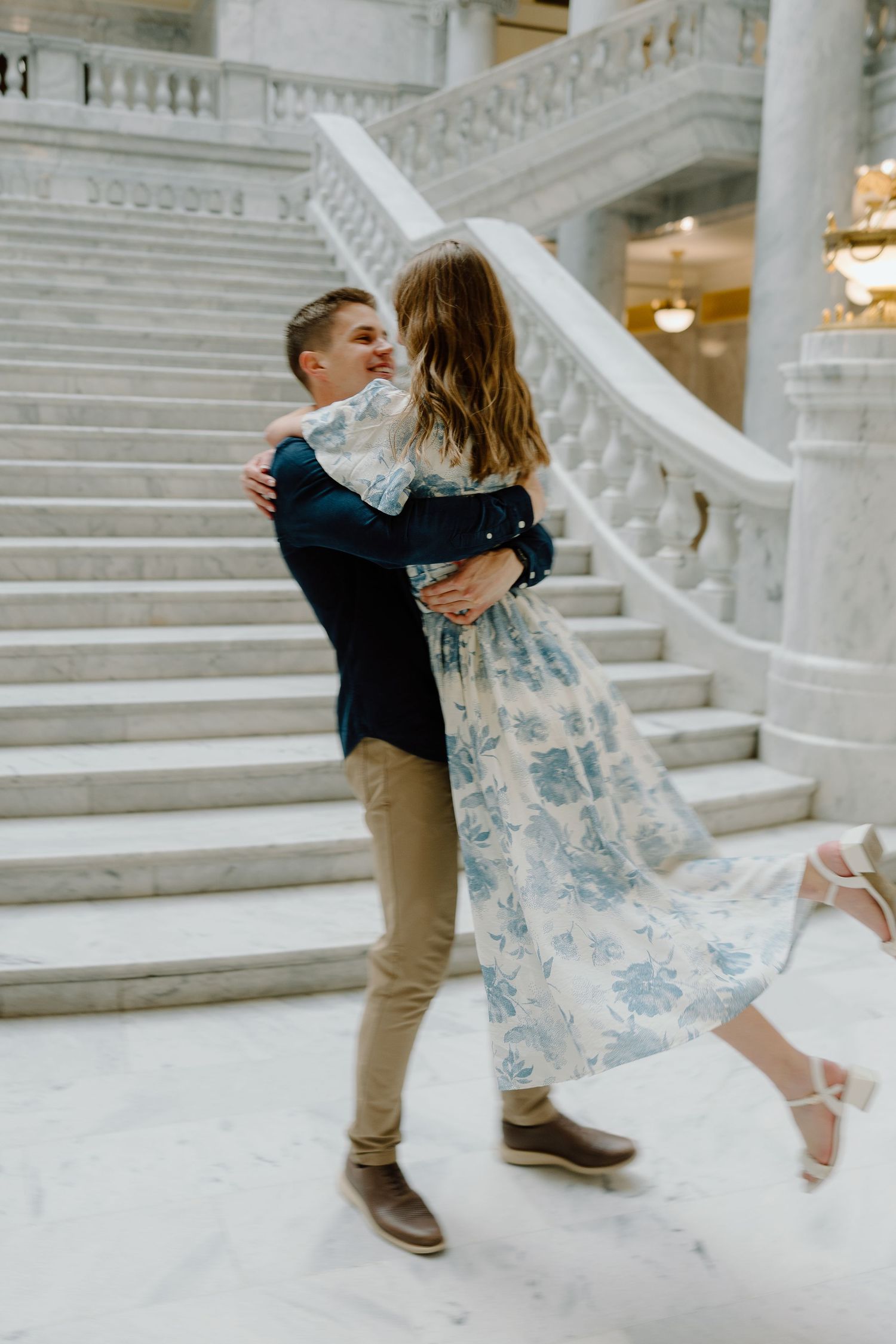 Couple dancing and embracing joyfully at the base of ornate marble staircase indoors.