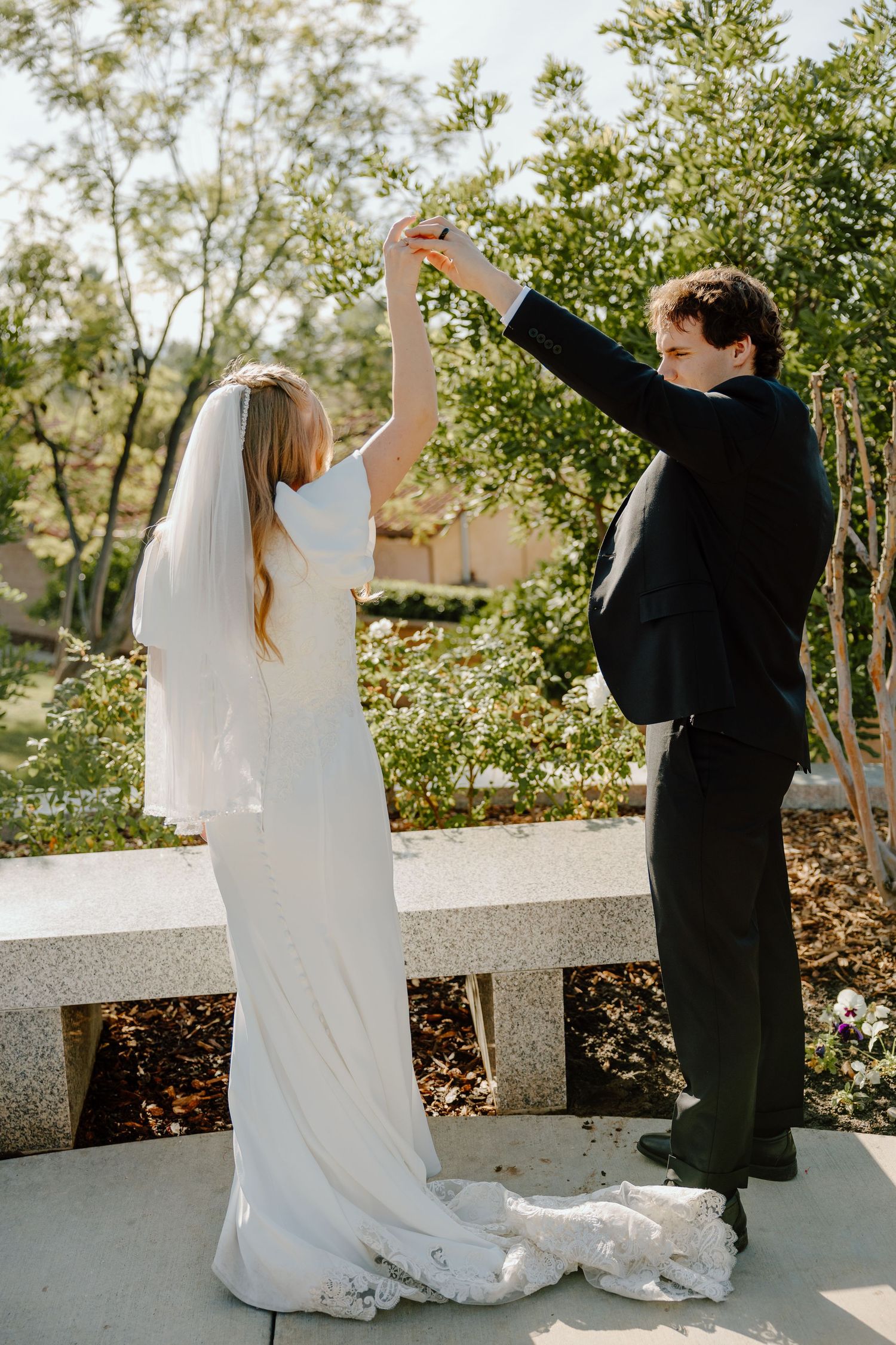 Bride and groom dancing outdoors, bride in white gown with veil, groom in dark suit, surrounded by lush greenery.