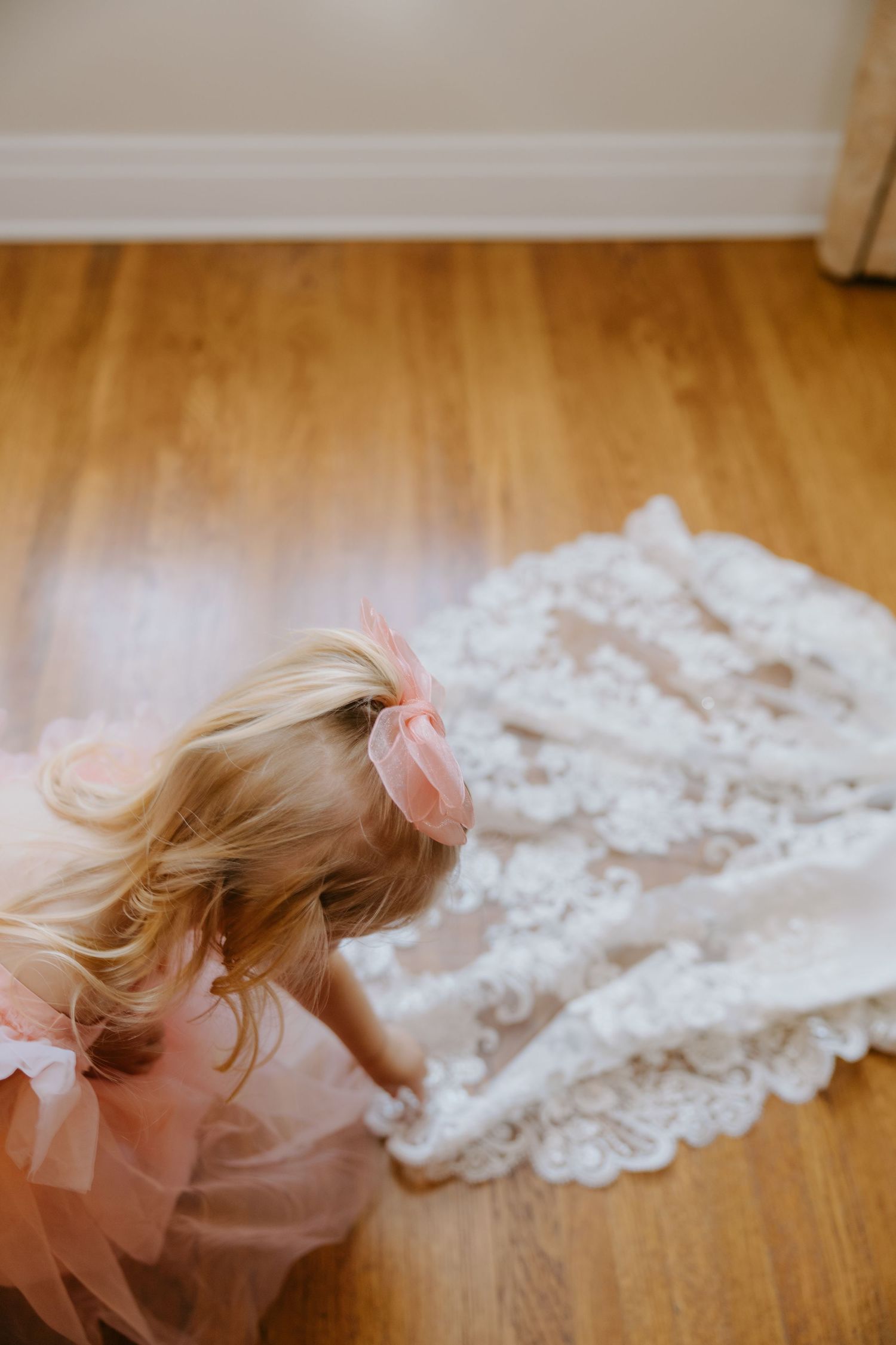 Young flower girl in pink tutu dress touching lace wedding dress train on hardwood floor.