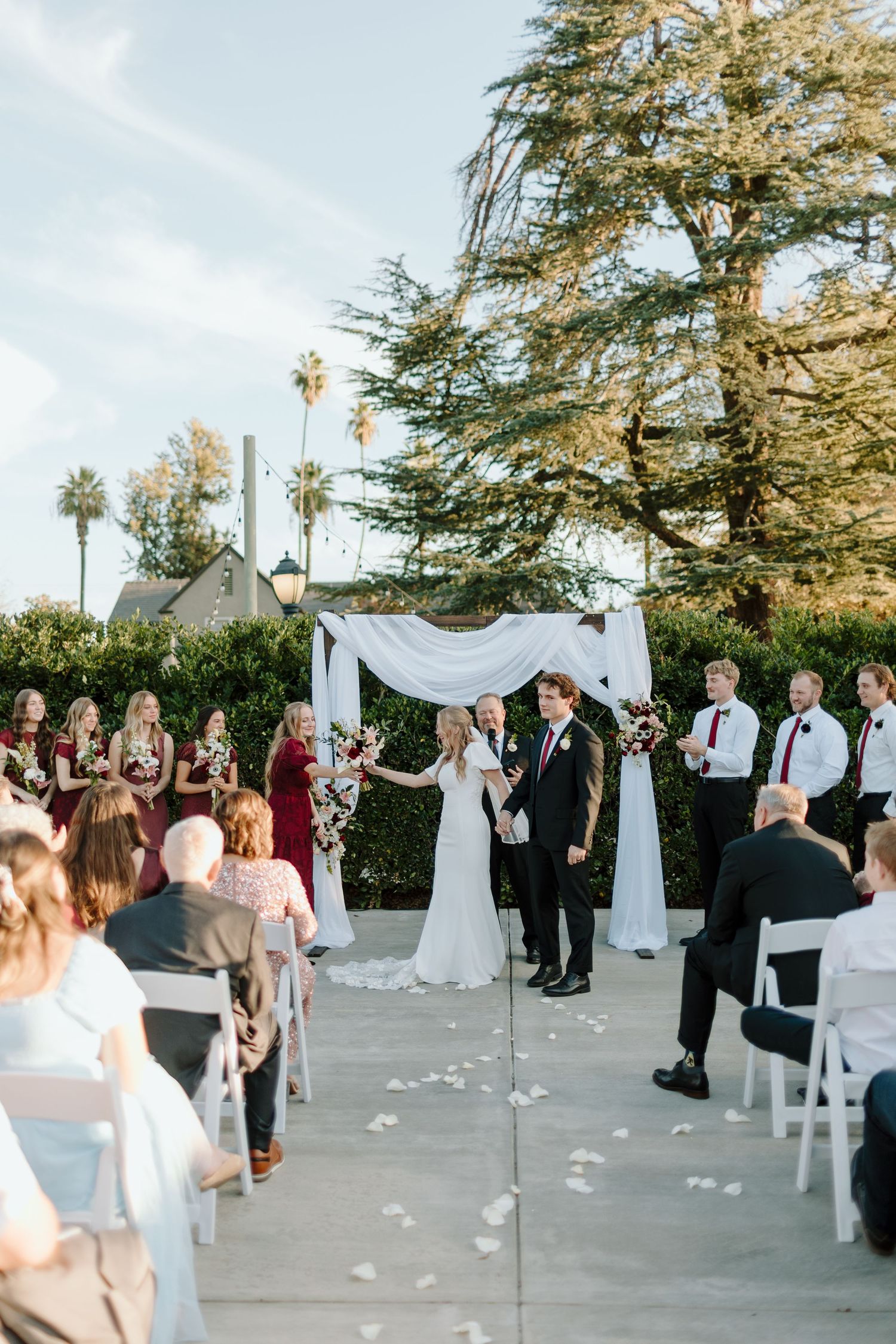 Outdoor wedding ceremony with bride and groom at floral arch, bridesmaids in burgundy, groomsmen in black, surrounded by lush greenery.