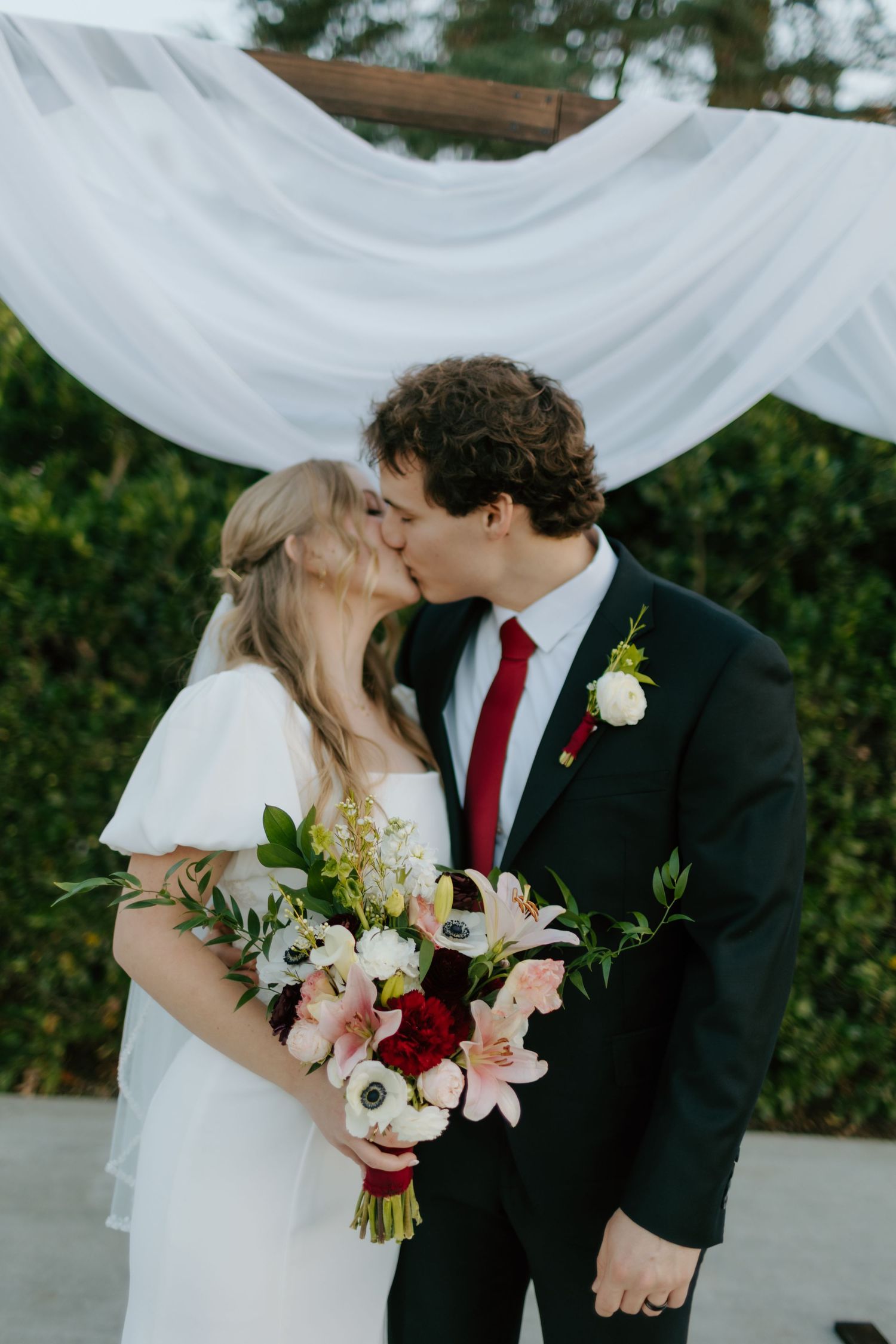 Newlyweds share their first kiss under a white draped arch, bride holding a pink and white floral bouquet.