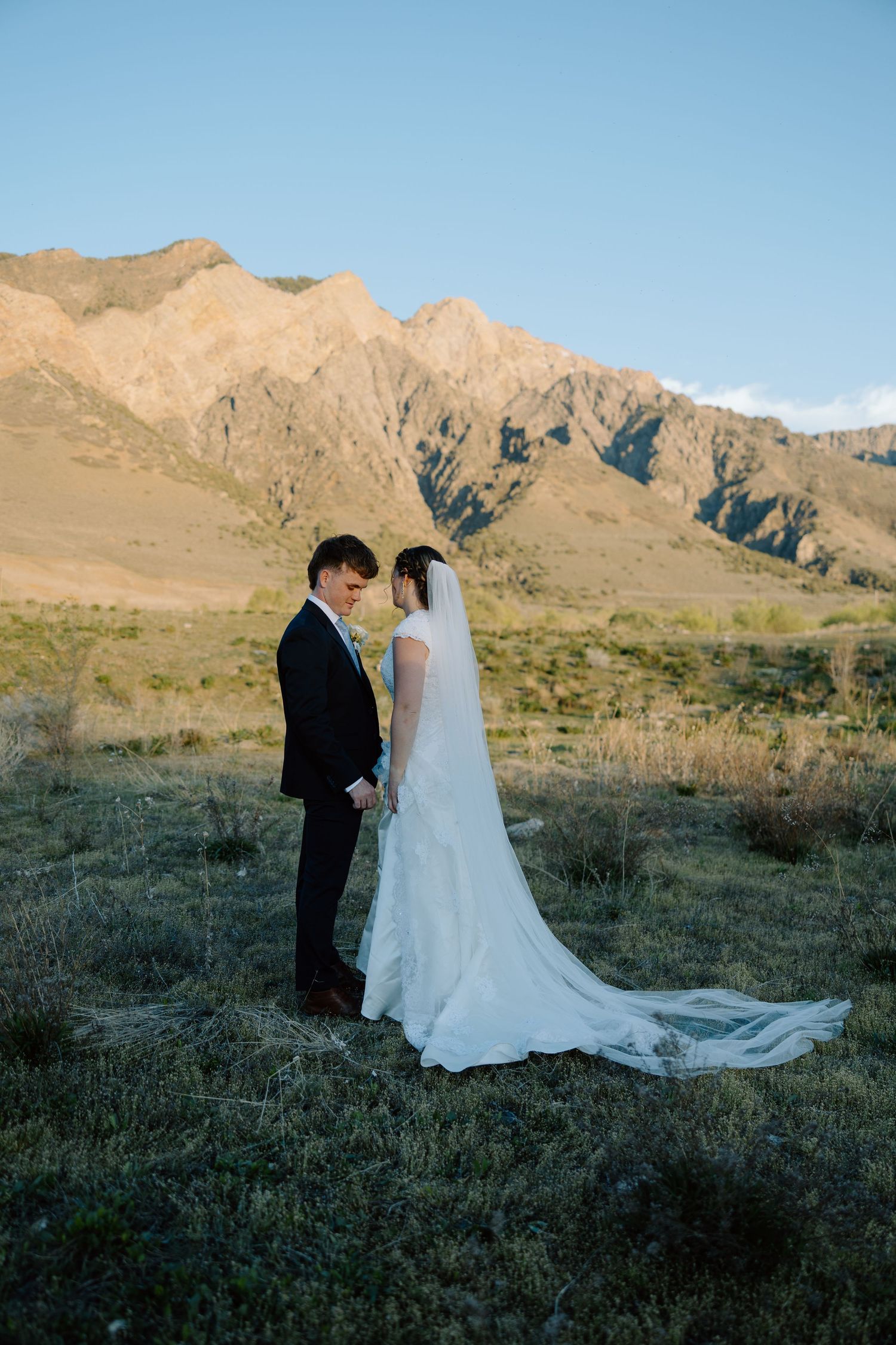 Newlyweds hold hands facing each other in a sunlit mountain meadow, brides veil trailing behind.