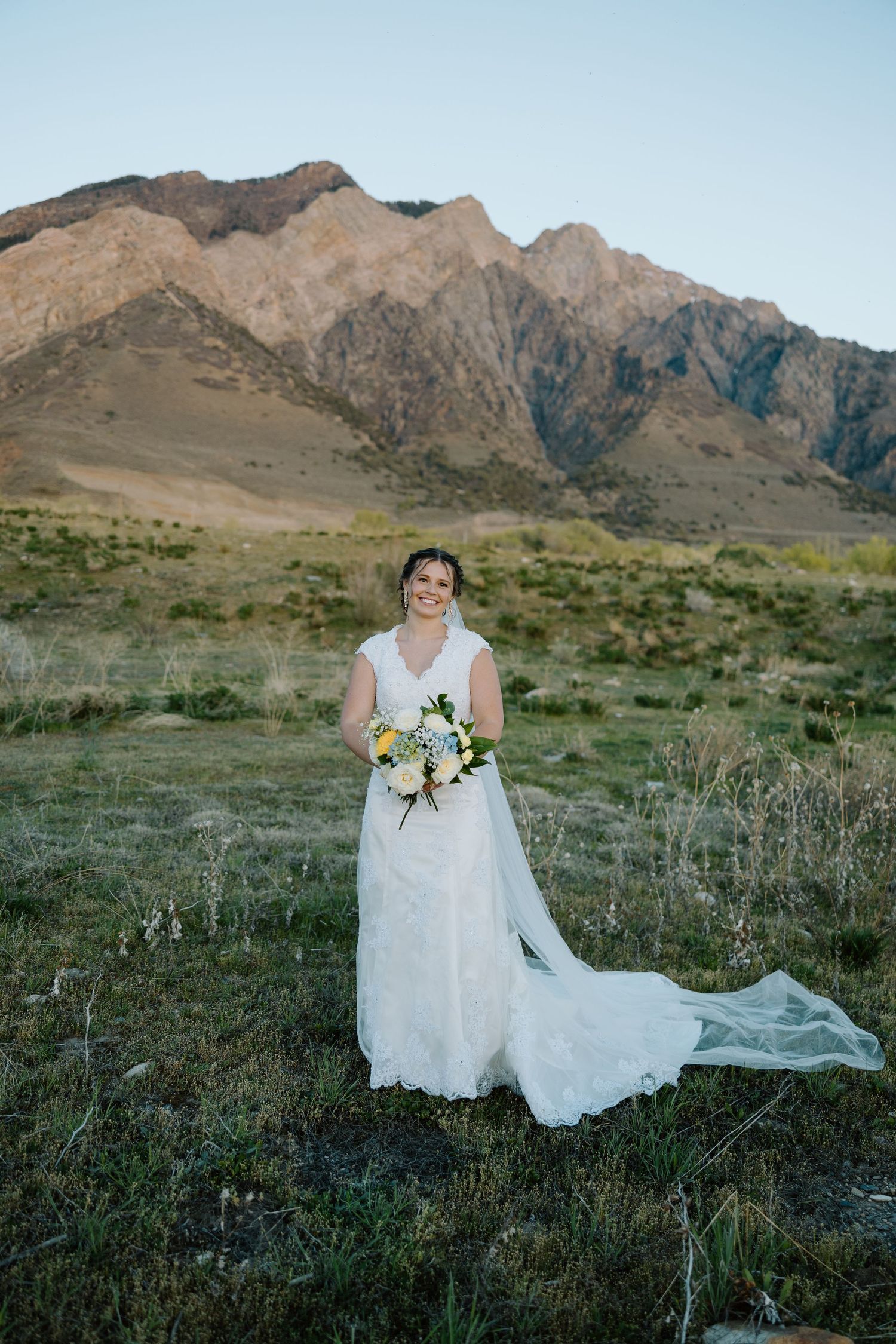 Radiant bride in lace gown holds bouquet while posing alone in a field with stunning mountain scenery.