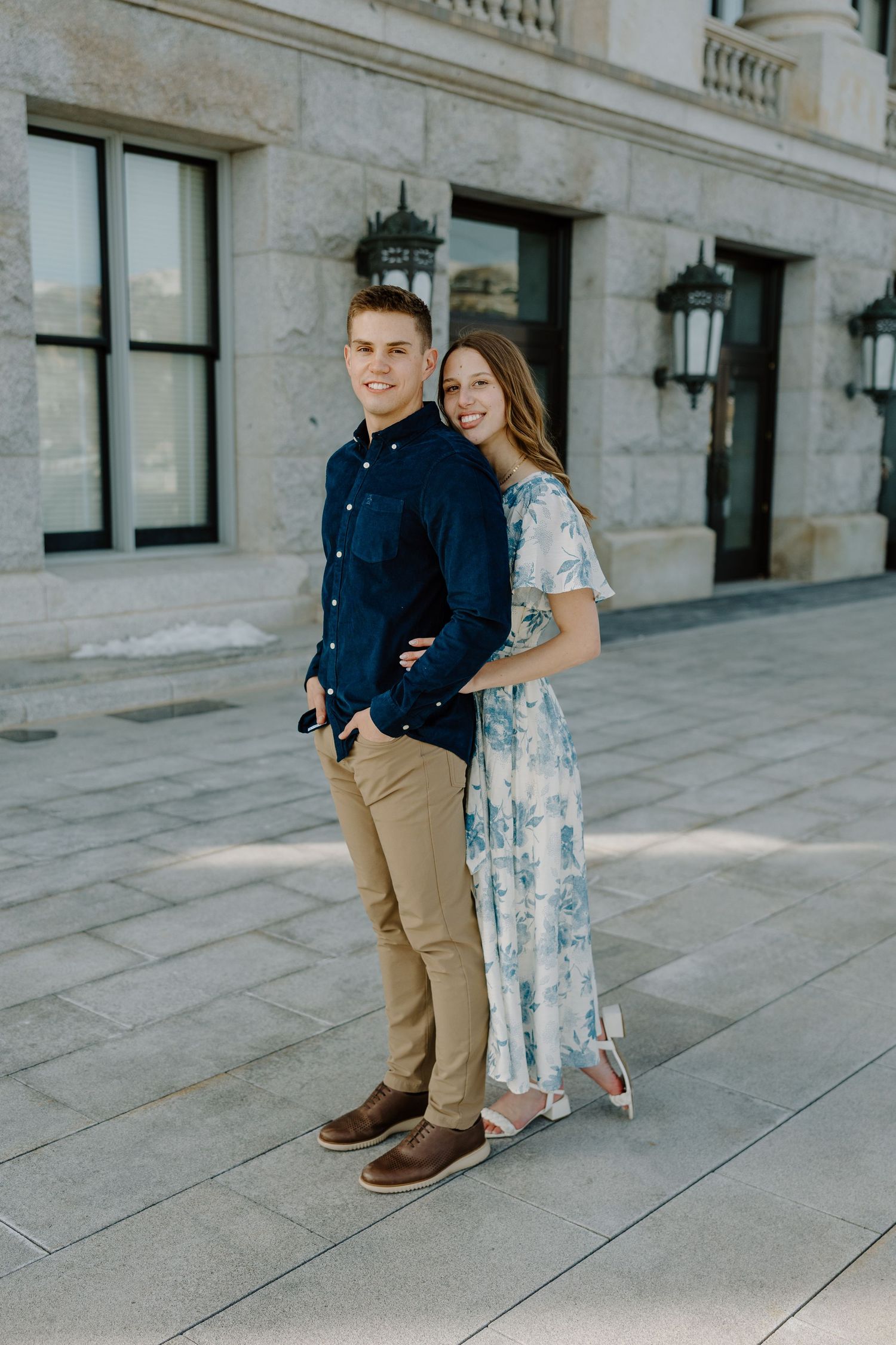 Smiling couple posing together outside classic stone building, man in navy shirt, woman in floral maxi dress.