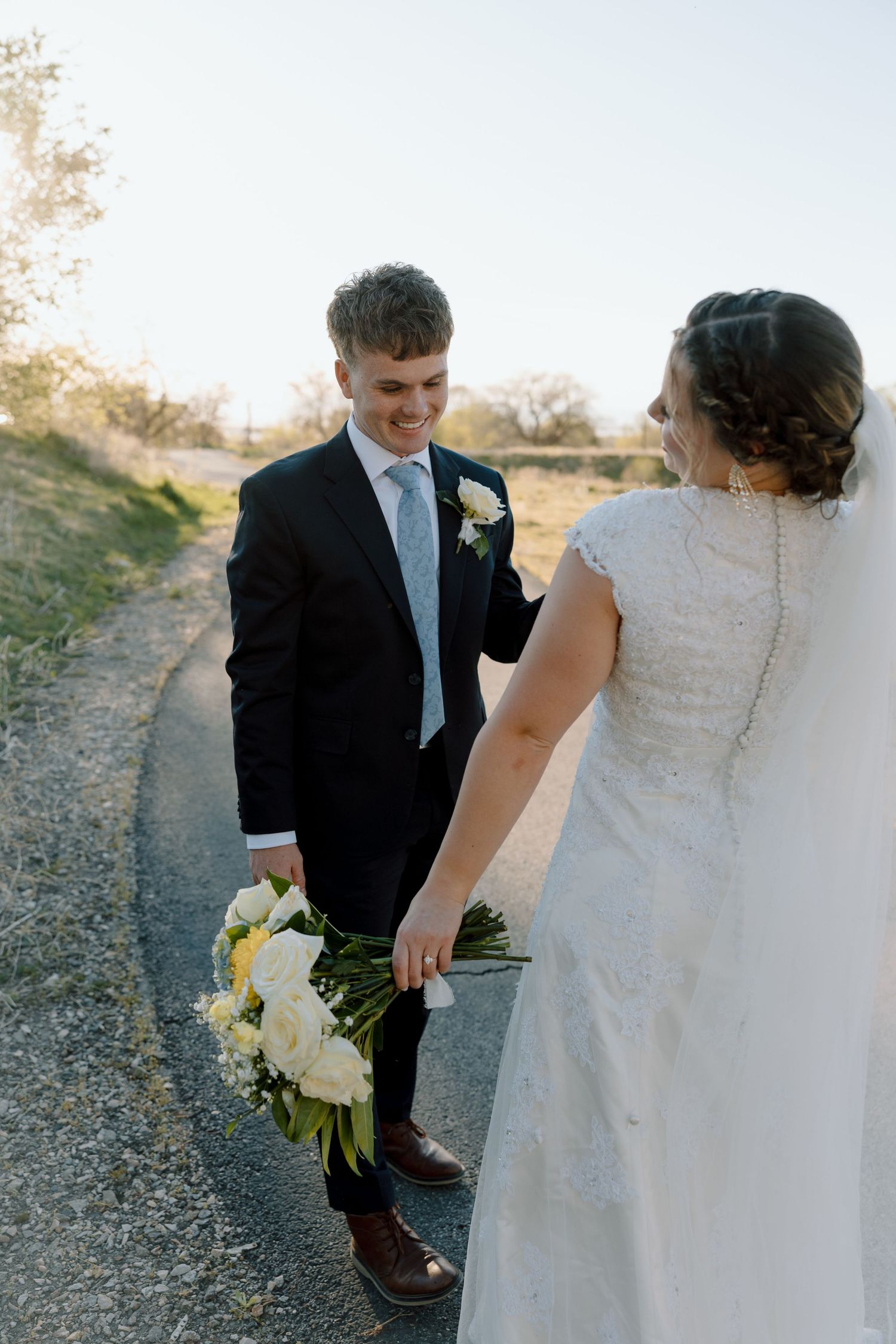 Groom smiles at bride while holding white floral bouquet on a scenic outdoor path.