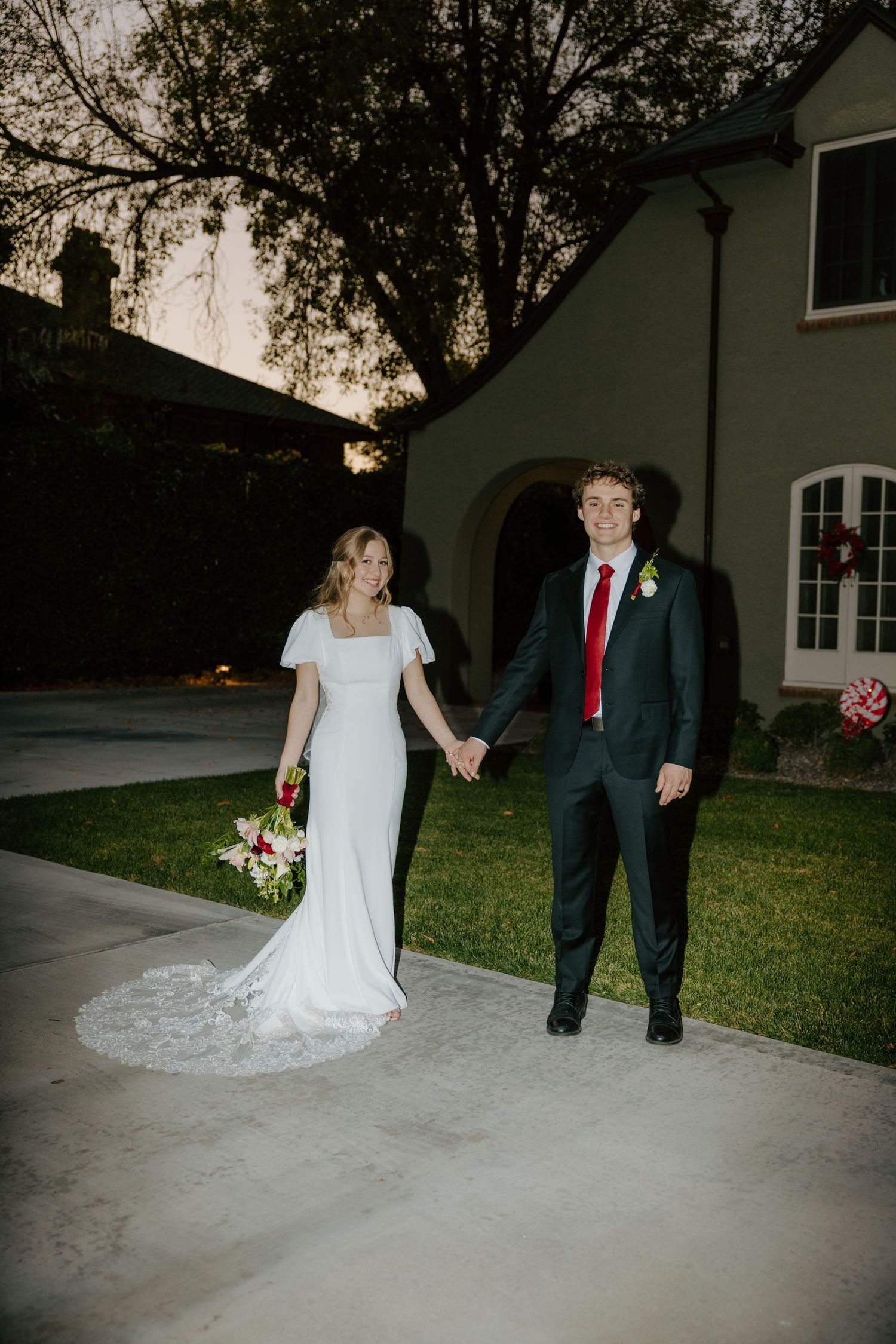 Bride in white gown and groom in dark suit with red tie hold hands outside elegant home at dusk.
