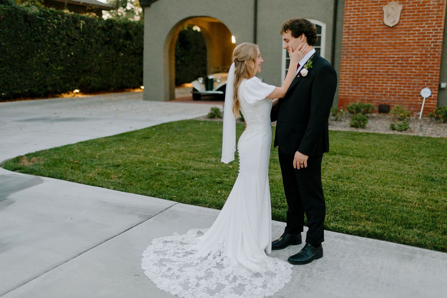 Bride and groom share a first look moment outdoors, with groom adjusting bride's white lace-train wedding dress.