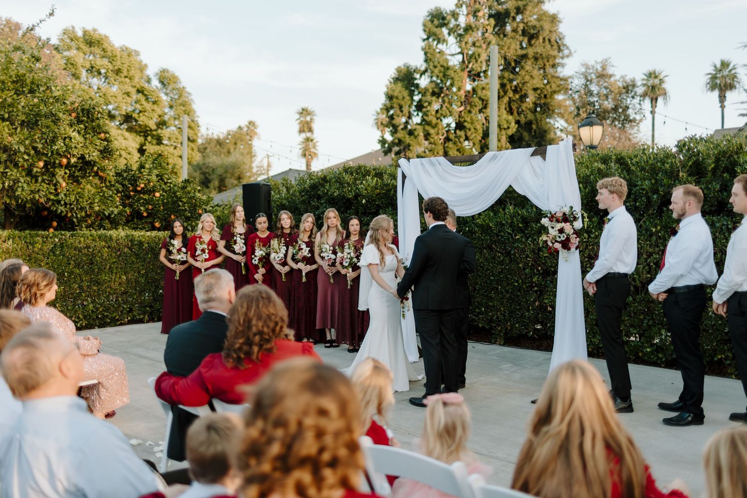 Outdoor wedding ceremony with bride, groom, wedding party in burgundy dresses and white shirts under a draped arch.