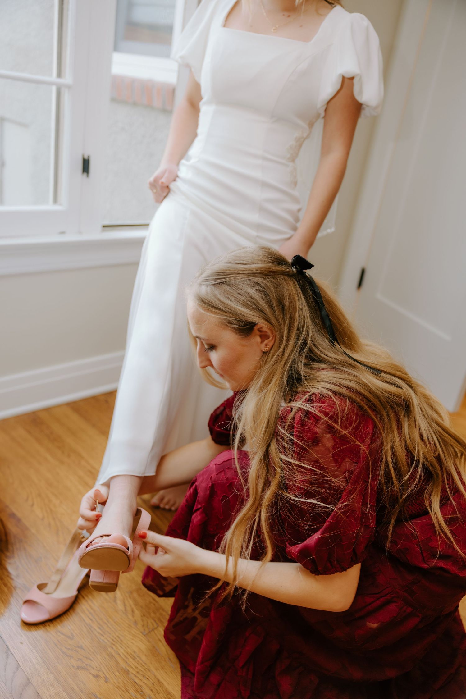 Bridesmaid in burgundy dress kneels to help bride put on blush heels before the wedding ceremony.