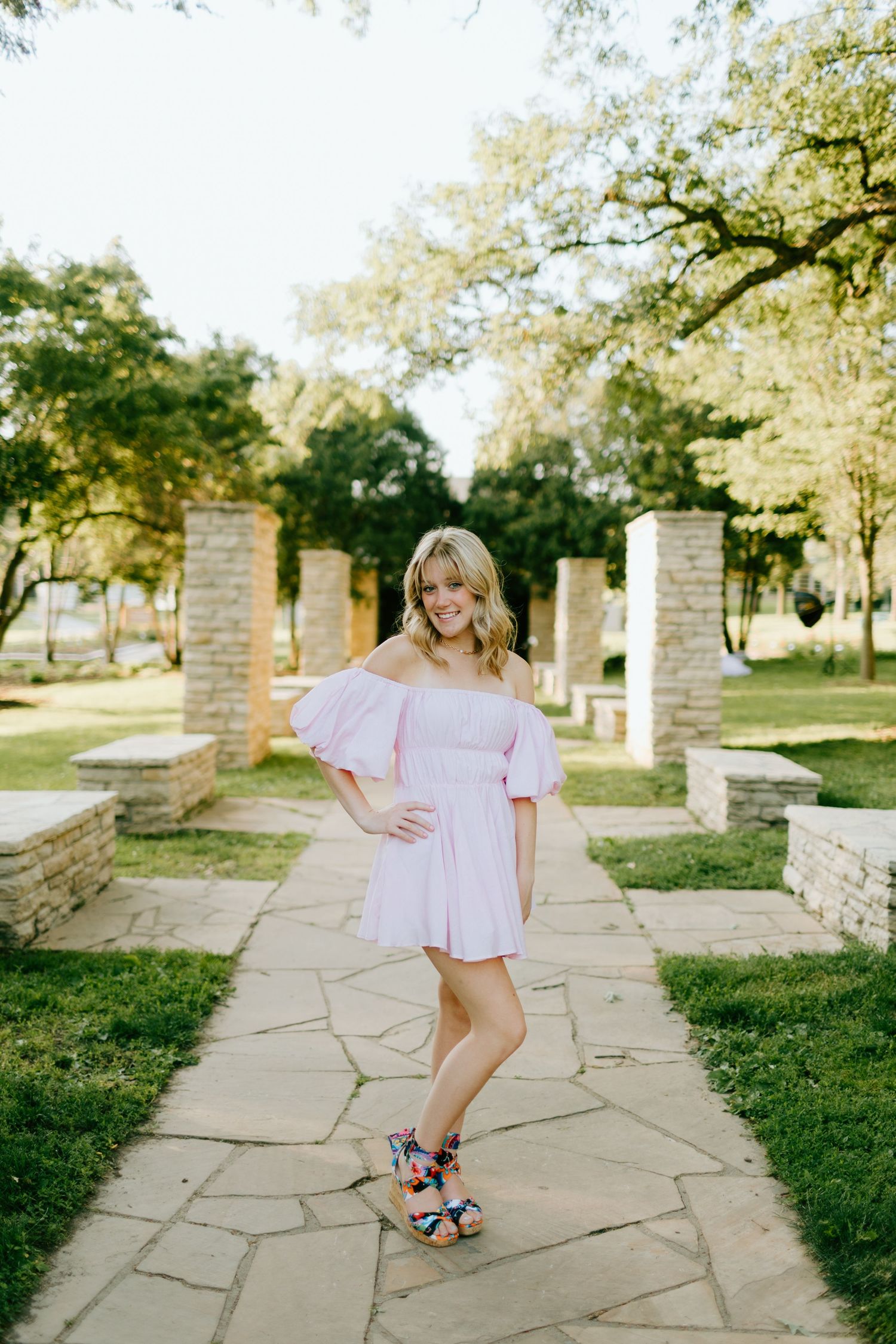 Young woman in pink off-shoulder mini dress posing on stone pathway in lush garden park.