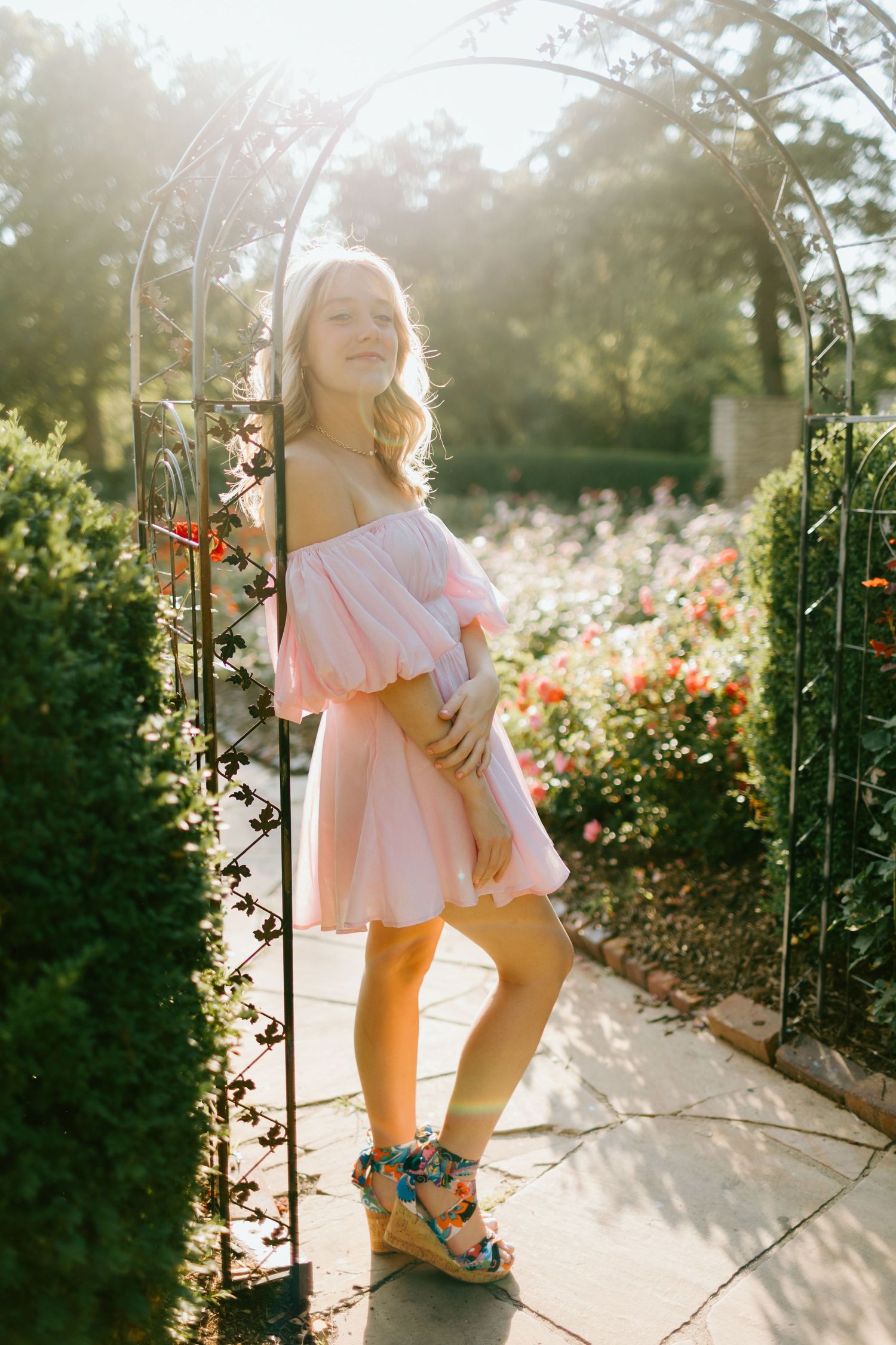 Blonde woman in pink off-shoulder dress posing under garden arch with roses in golden sunlight.