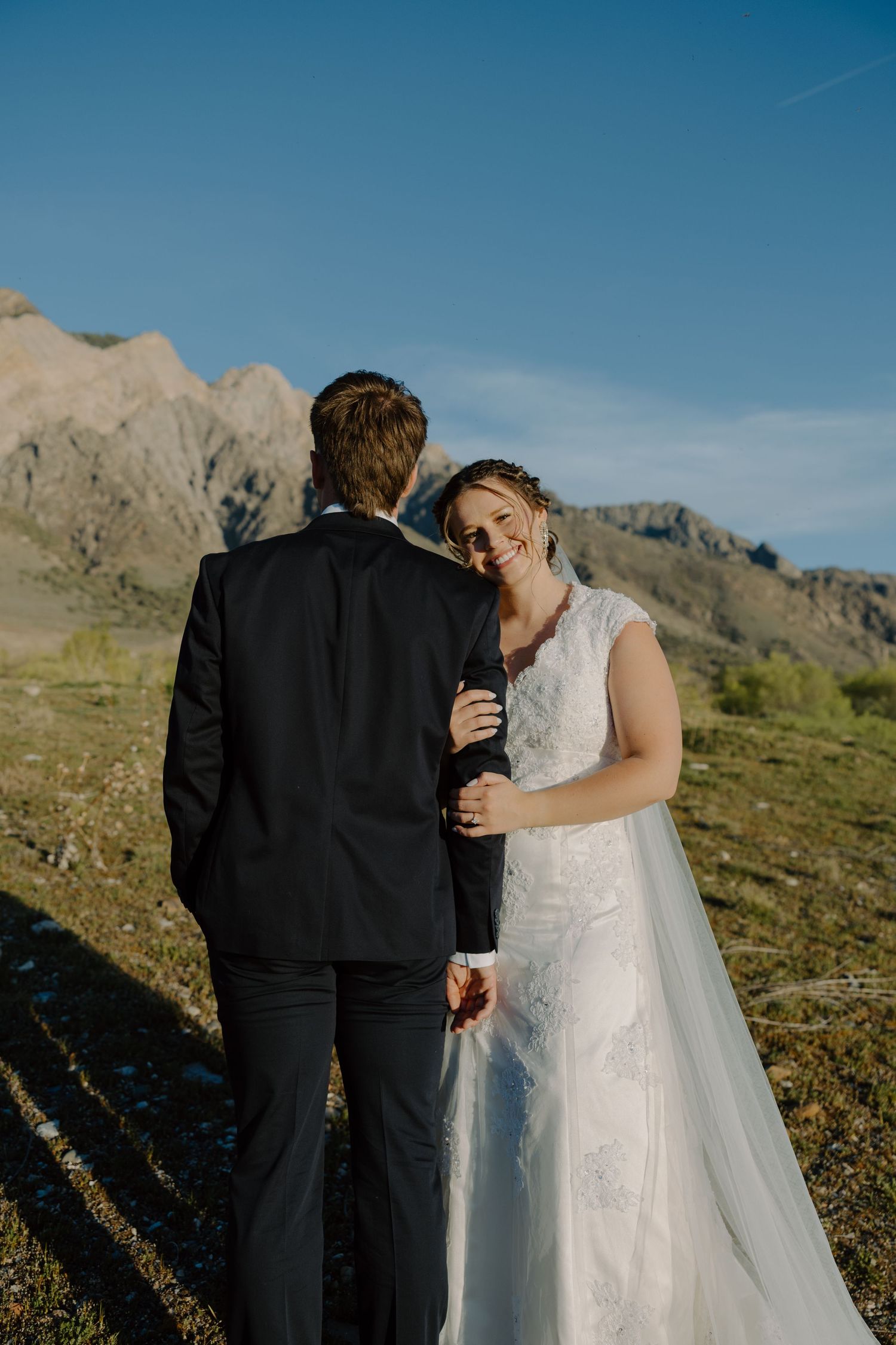 Radiant bride smiles over shoulder while holding groom's hand in a mountain meadow.