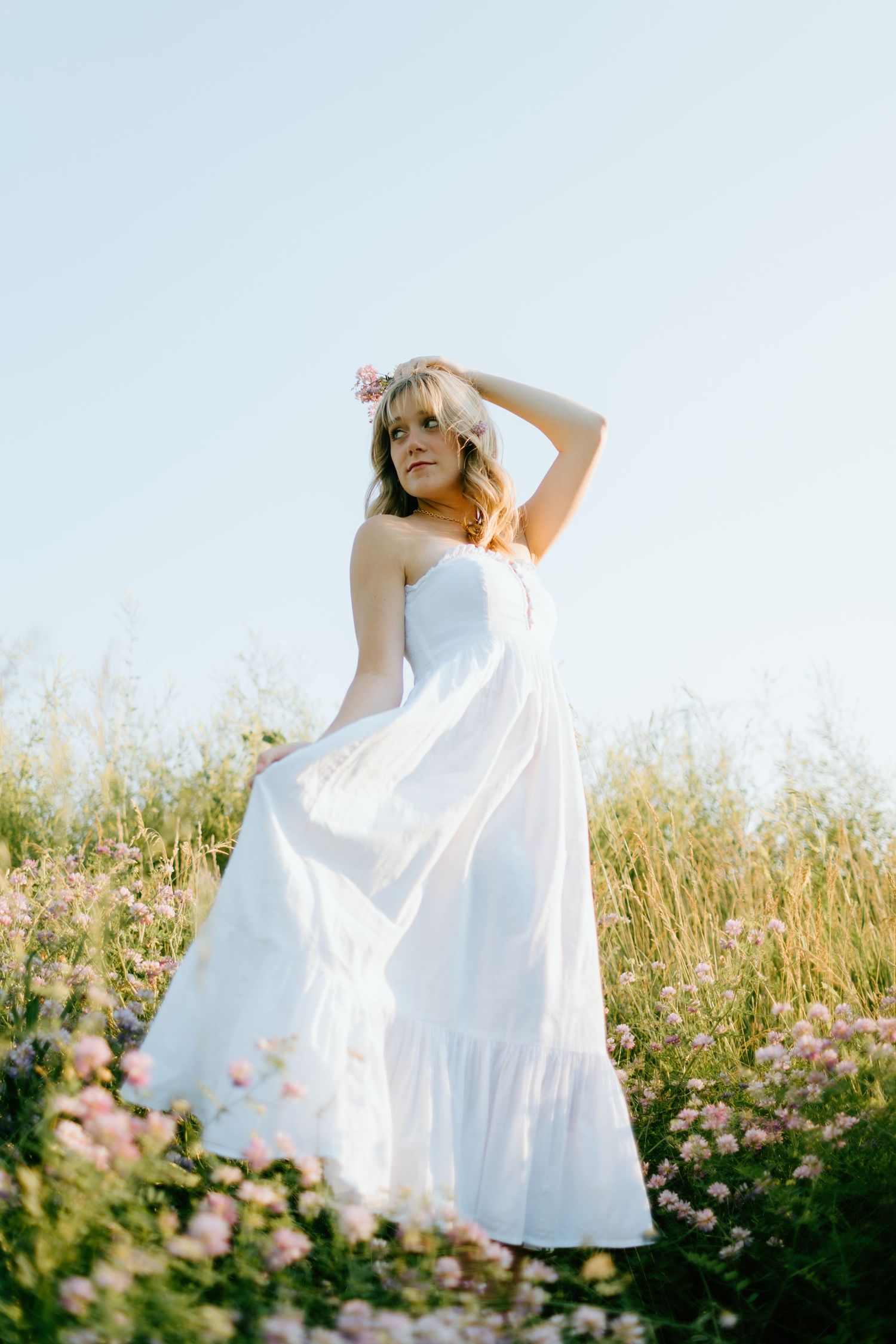 Woman in white maxi dress with flower crown standing in a wildflower meadow under a clear blue sky.