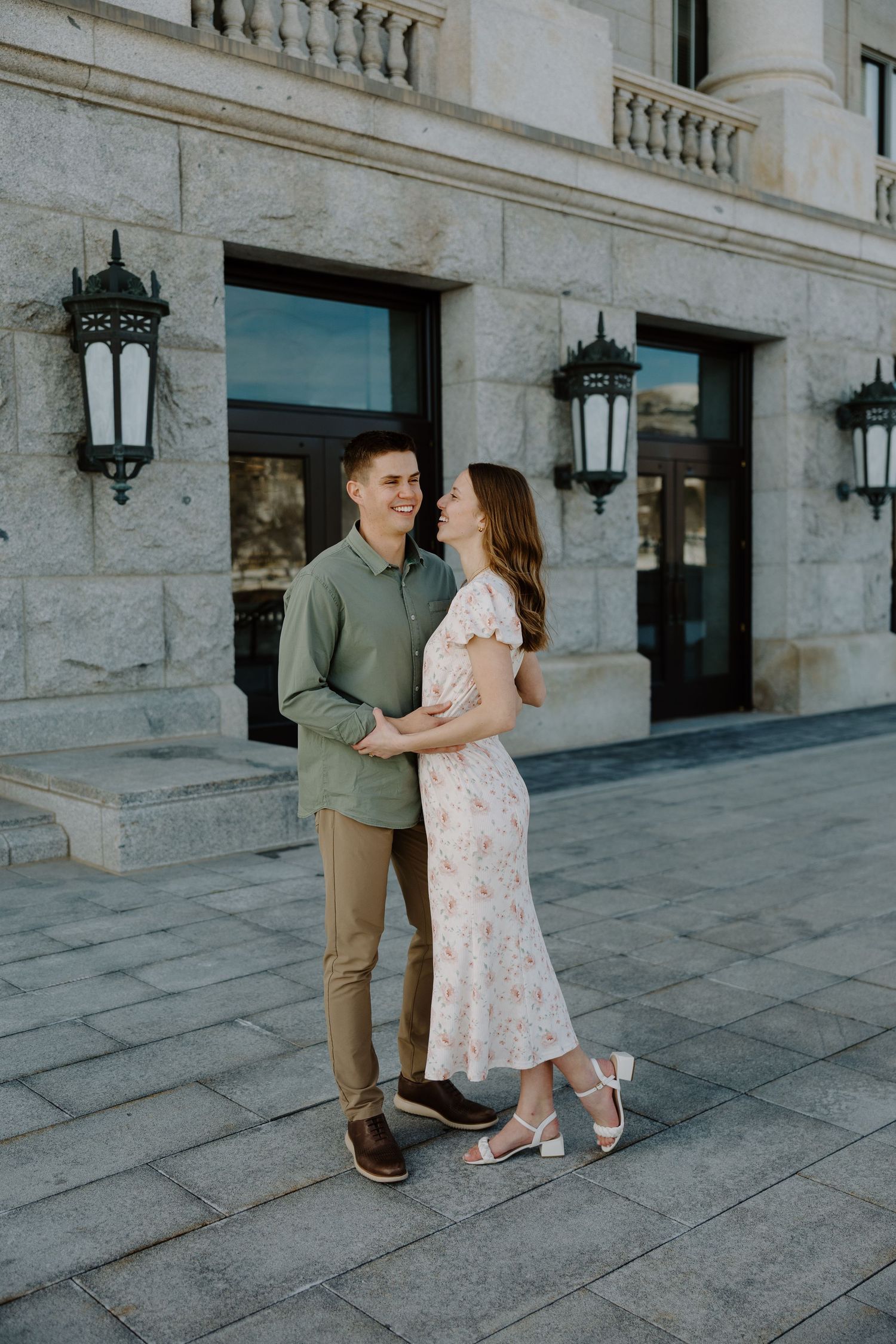 A smiling couple embraces outdoors in front of an elegant stone building, woman in floral dress, man in green shirt.