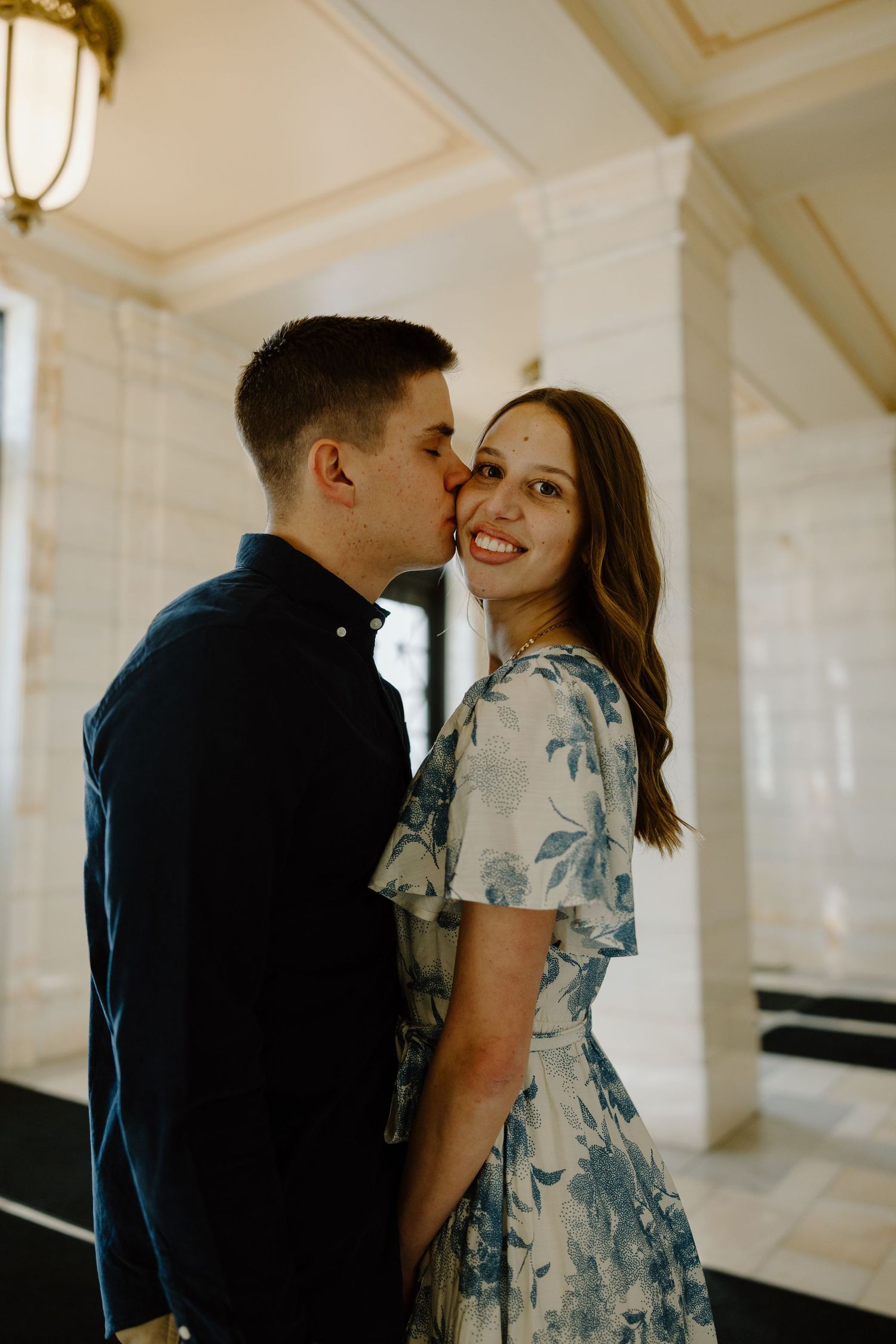 Couple sharing a romantic moment, man kissing woman's cheek as she smiles inside an elegant building.