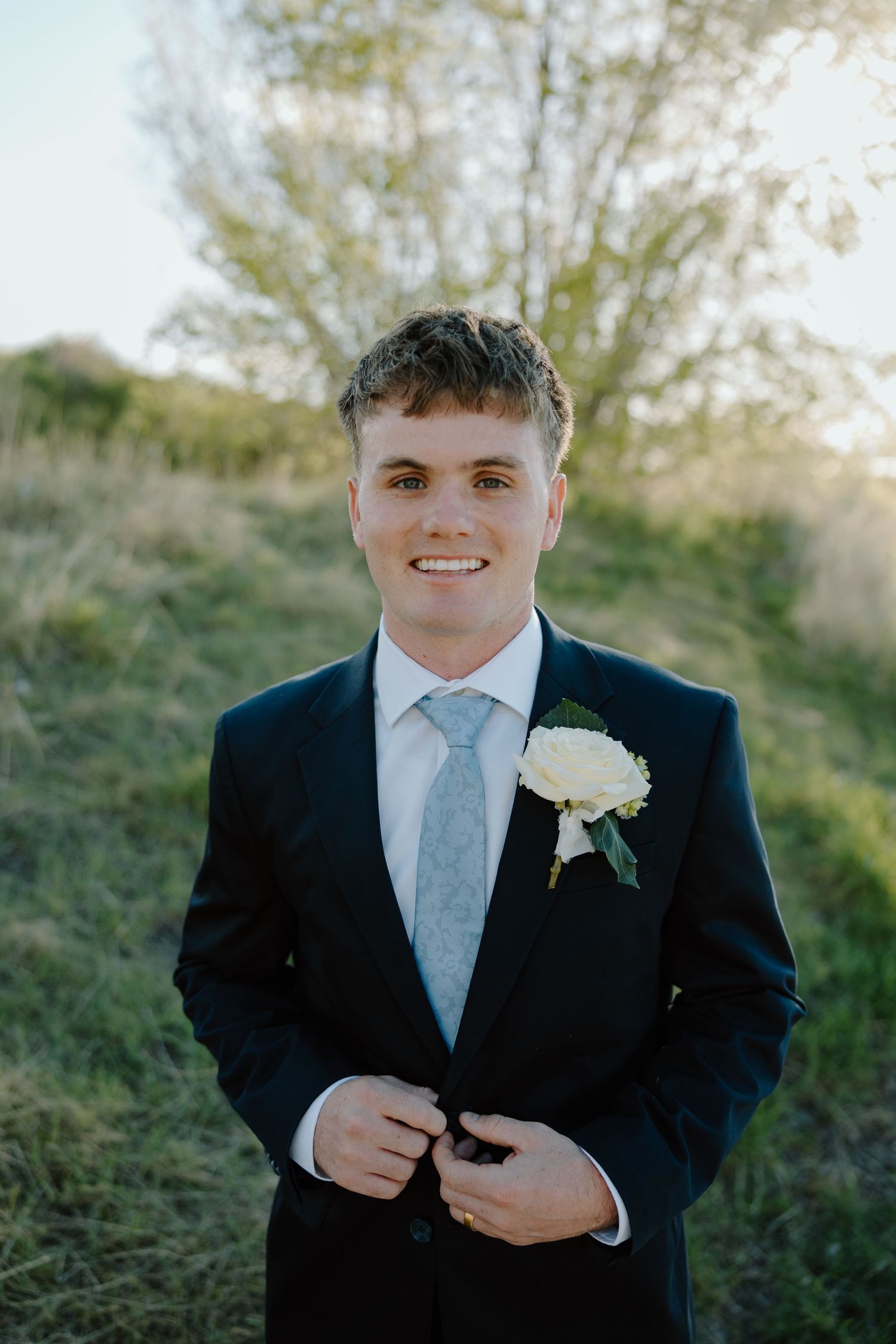Smiling groom in black suit with white boutonniere and light blue tie poses outdoors on wedding day.