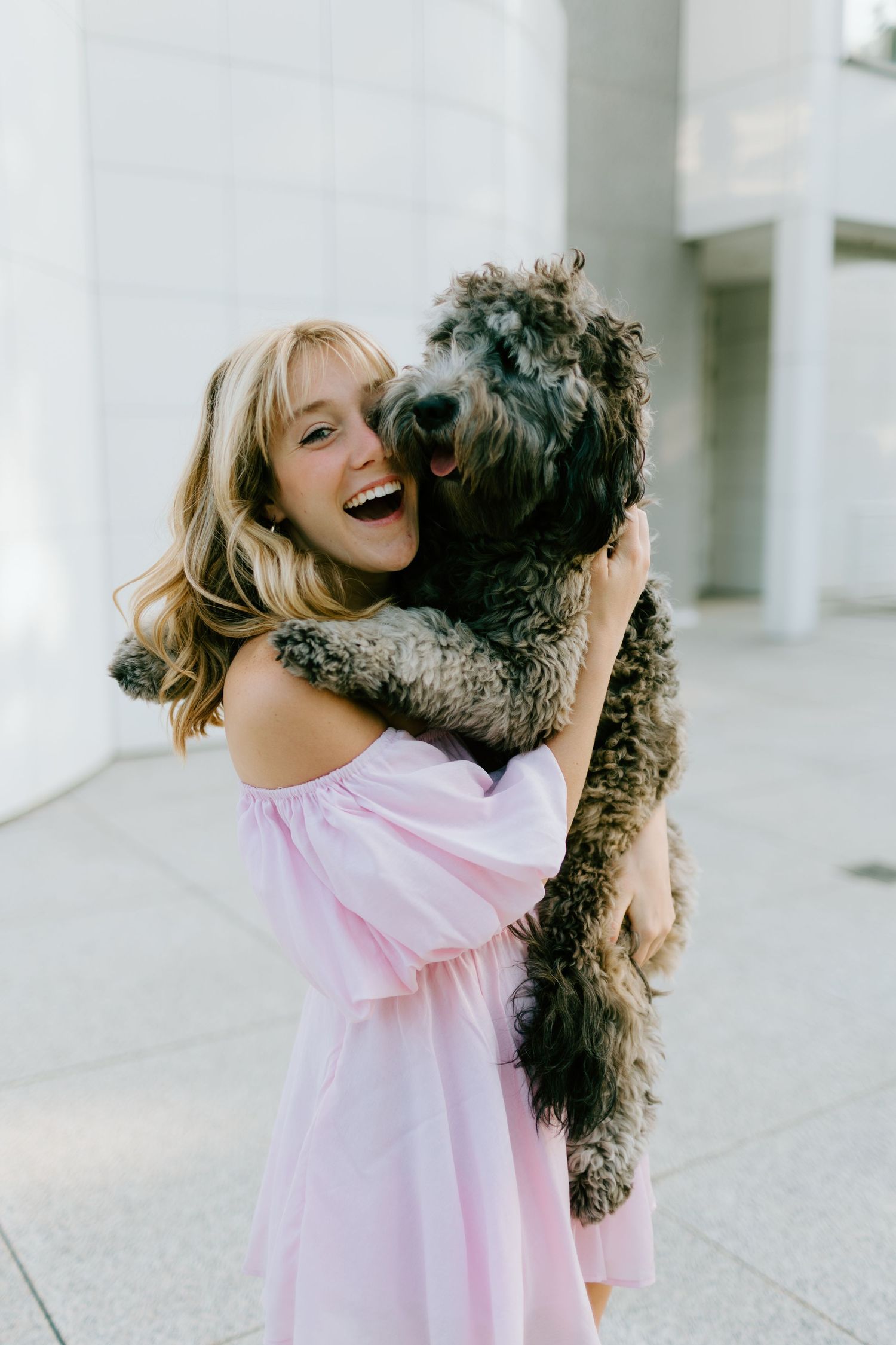 Laughing blonde woman in pink off-shoulder dress hugging a curly dark dog outdoors.