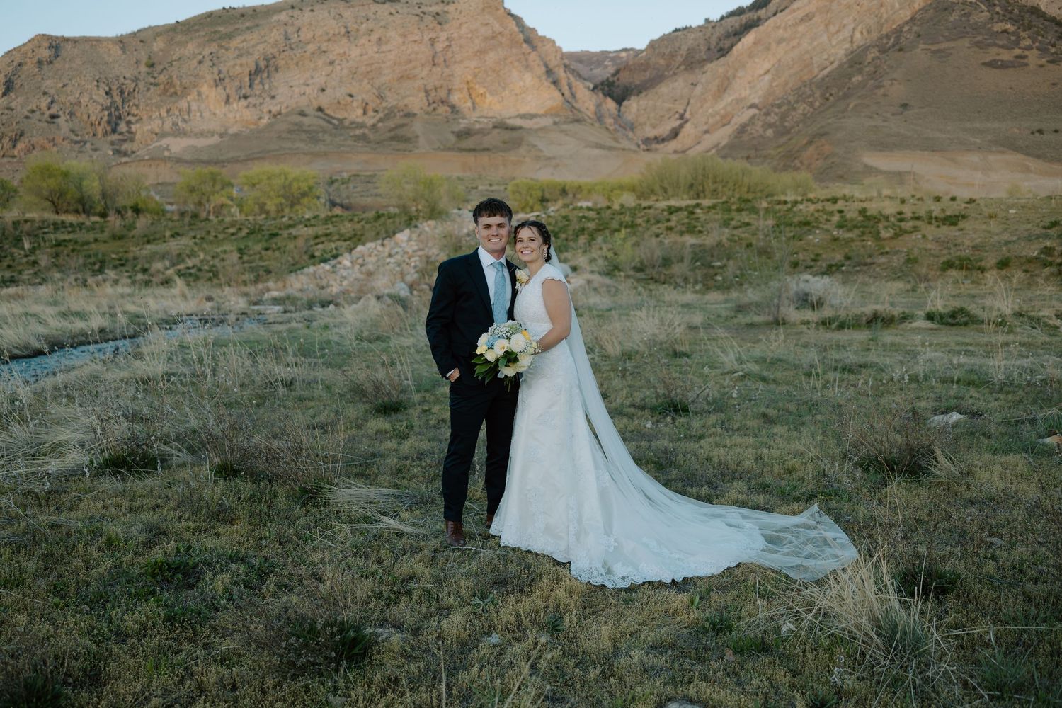 Bride and groom pose together in a scenic outdoor setting with mountain backdrop and golden grassy field.