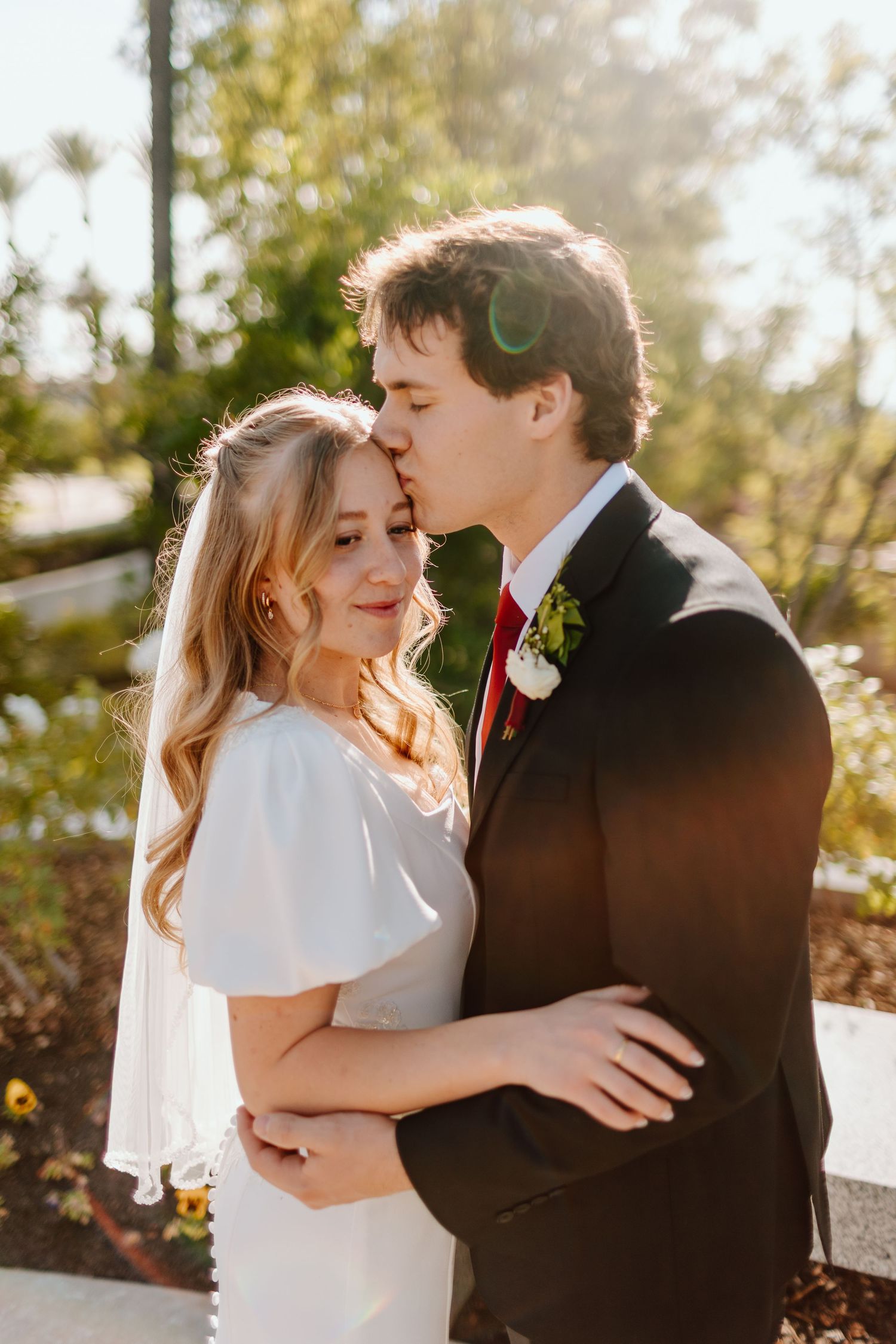 Groom kissing bride on forehead in golden sunlight, bride wearing veil.