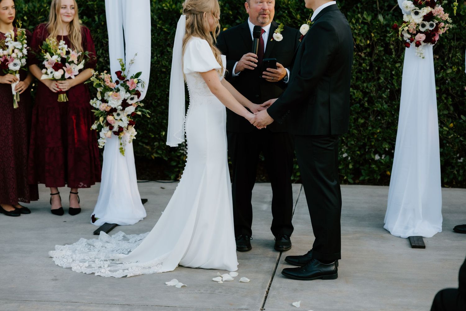 Bride and groom holding hands at outdoor wedding ceremony with bridesmaids in burgundy dresses.
