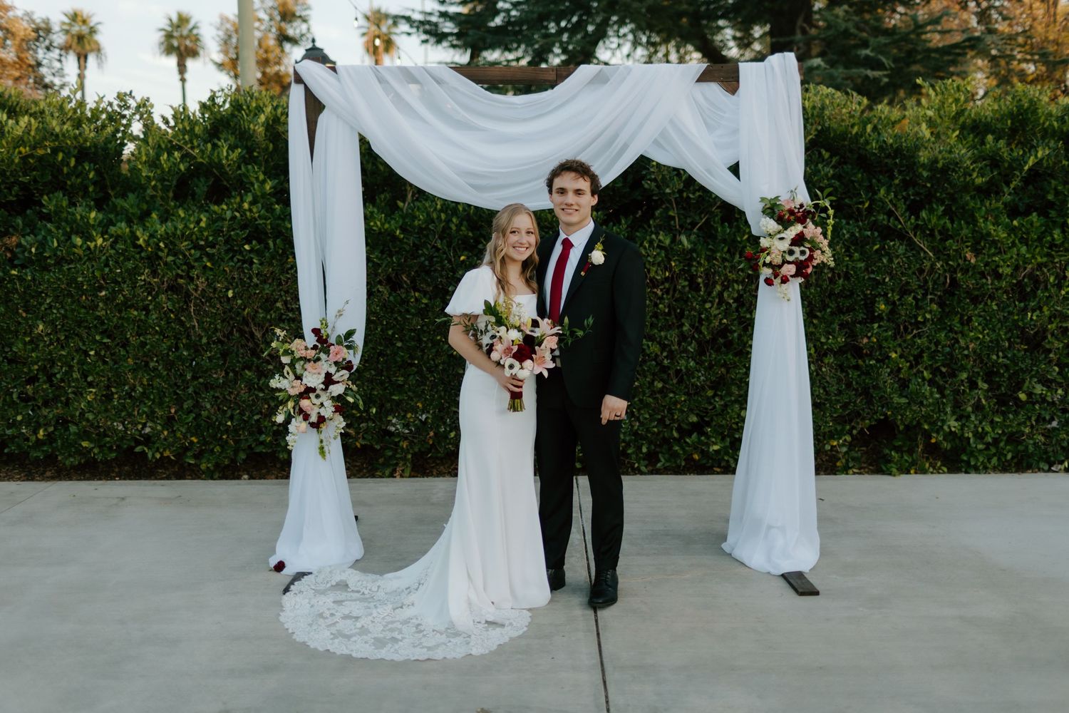 Bride and groom smiling under a white draped wedding arch adorned with floral arrangements outdoors.