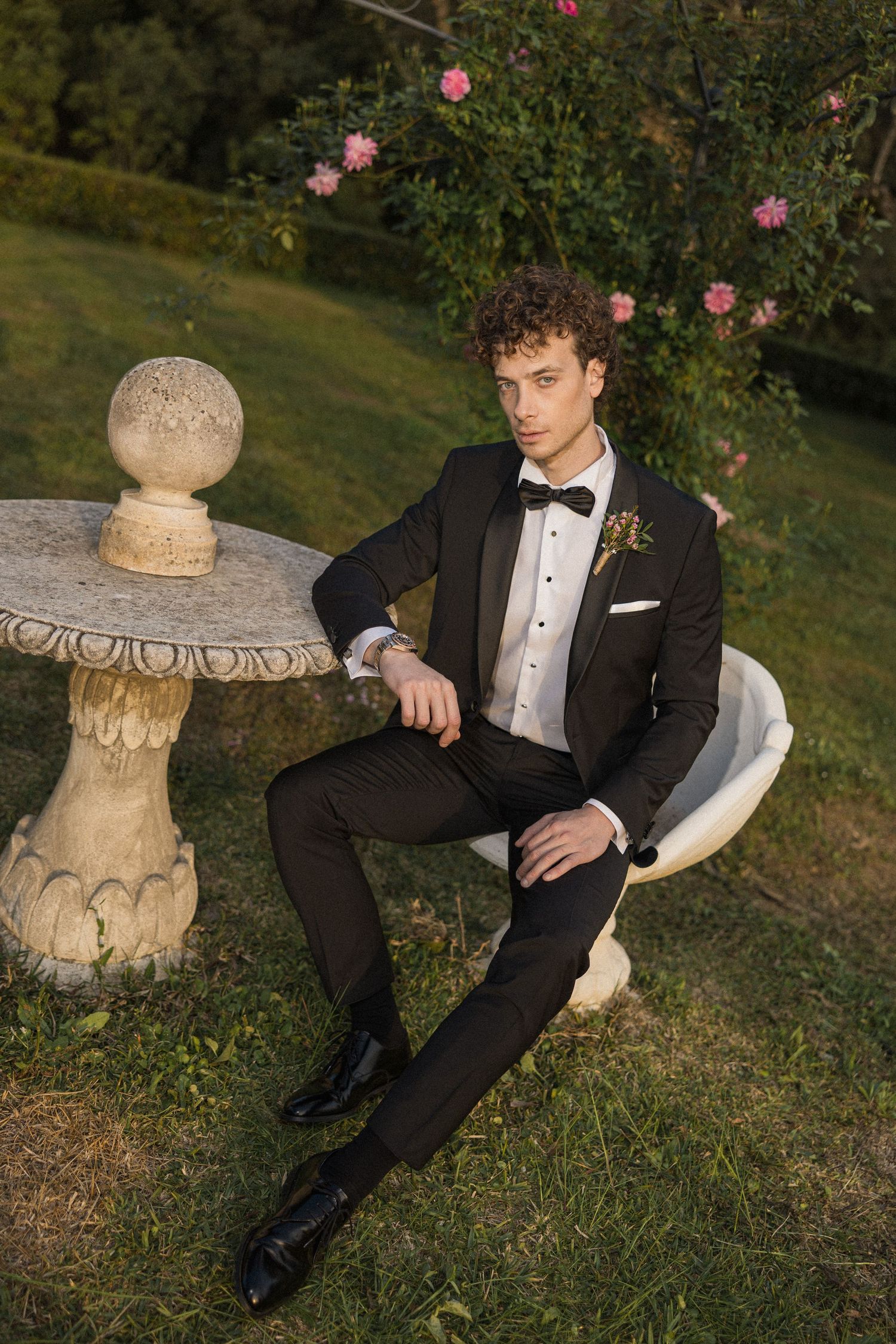 Classic black tuxedo with bow tie photographed in garden setting with ornate stone pedestal and pink roses in background.