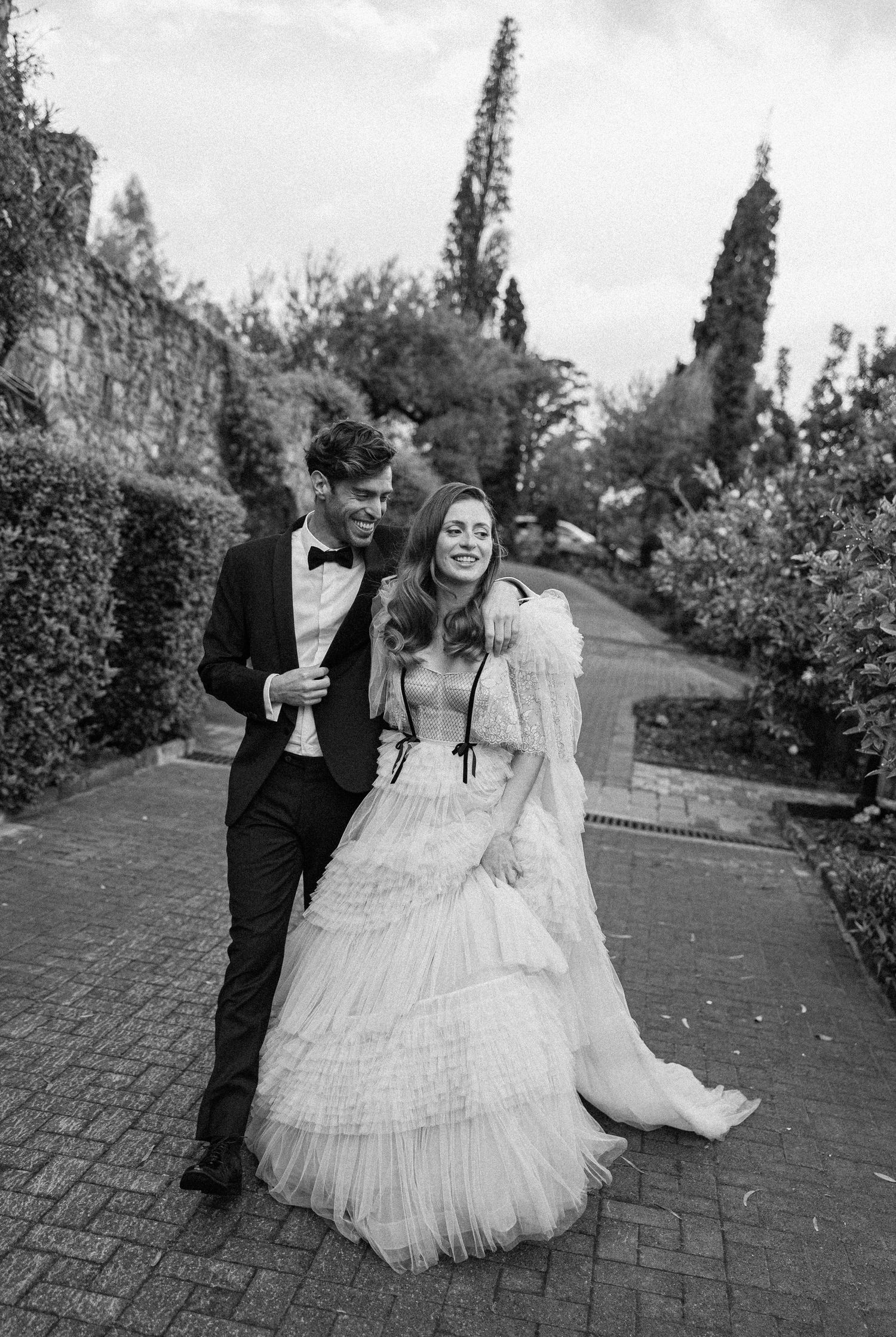A sequence of joyful moments captured at a wedding as people walk along a garden pathway in black and white.