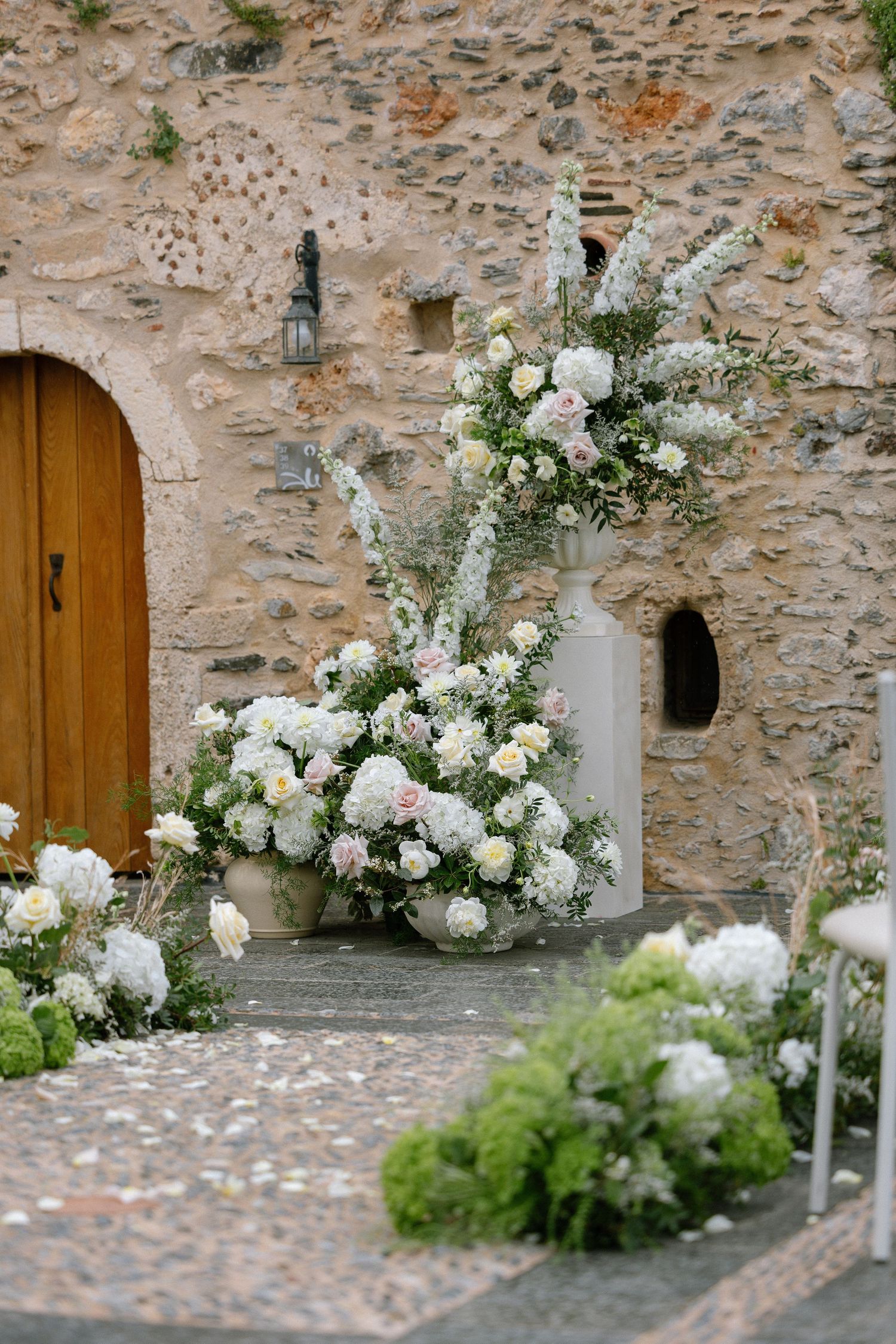 White floral arrangements with hydrangeas and greenery decorate a rustic stone wall with wooden doors.