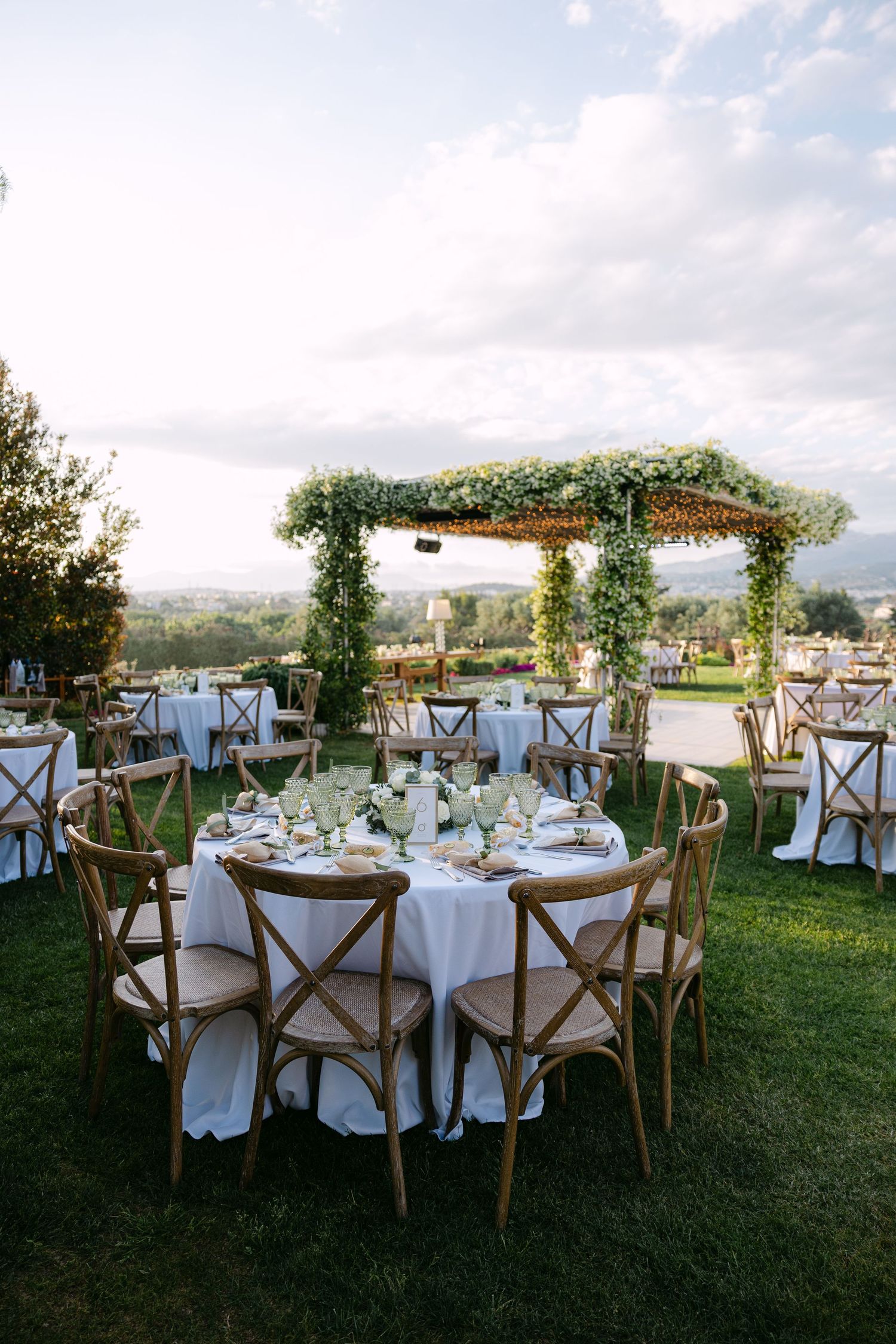 Elegant outdoor wedding reception setup with round tables, wooden cross-back chairs and a floral-covered pergola on a grassy lawn.