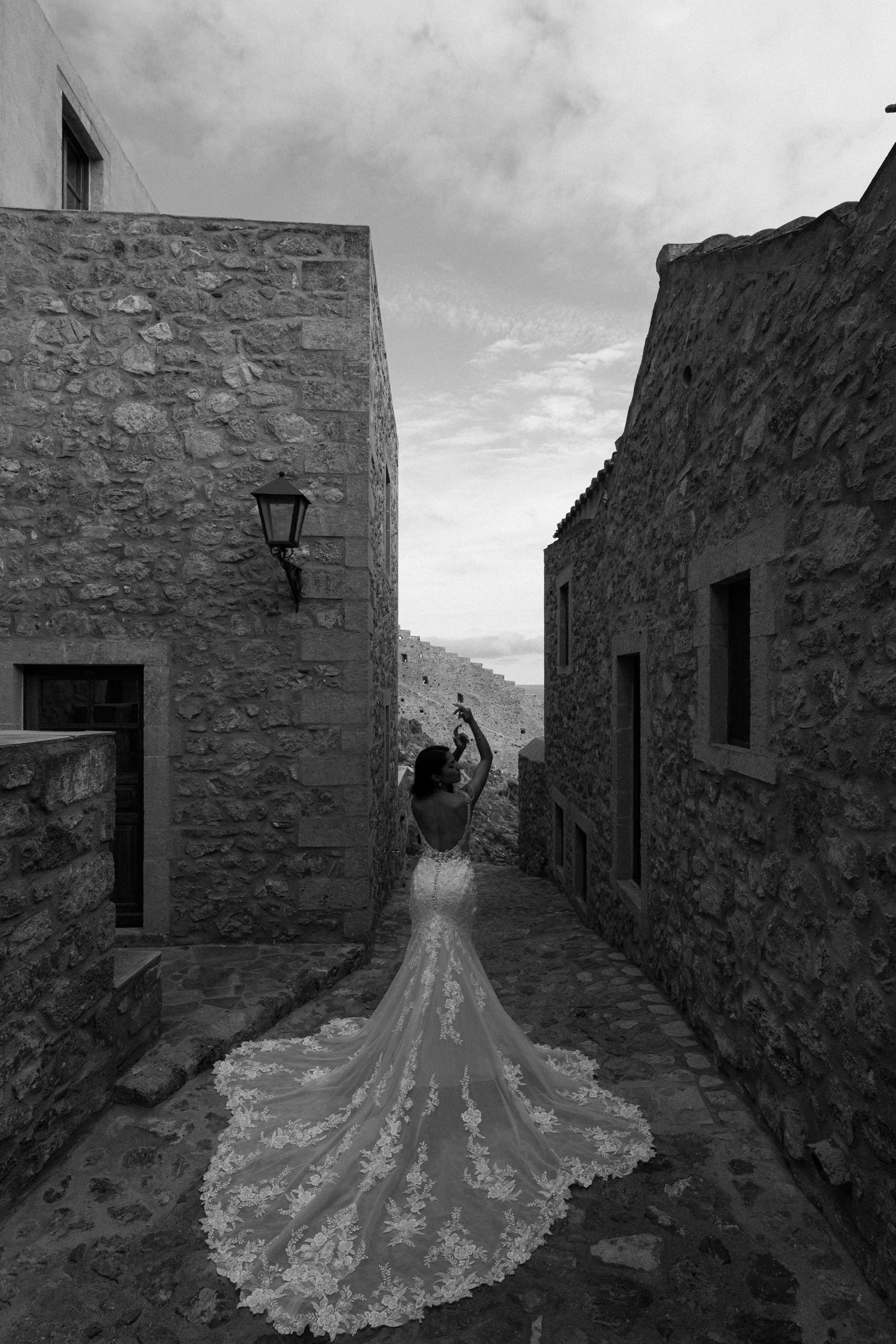 A dramatic black and white wedding dress silhouette with long lace train photographed in a narrow stone alleyway.