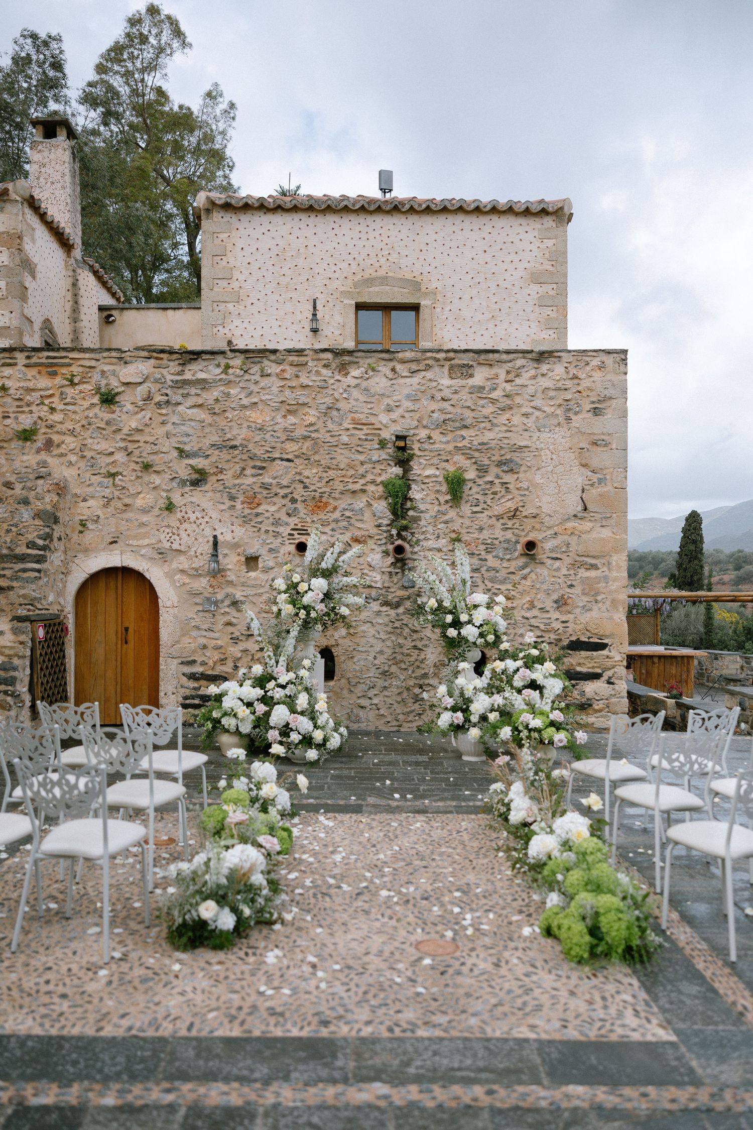 Rustic stone wall ceremony setup with white floral arrangements and modern chairs at an Italian villa courtyard.