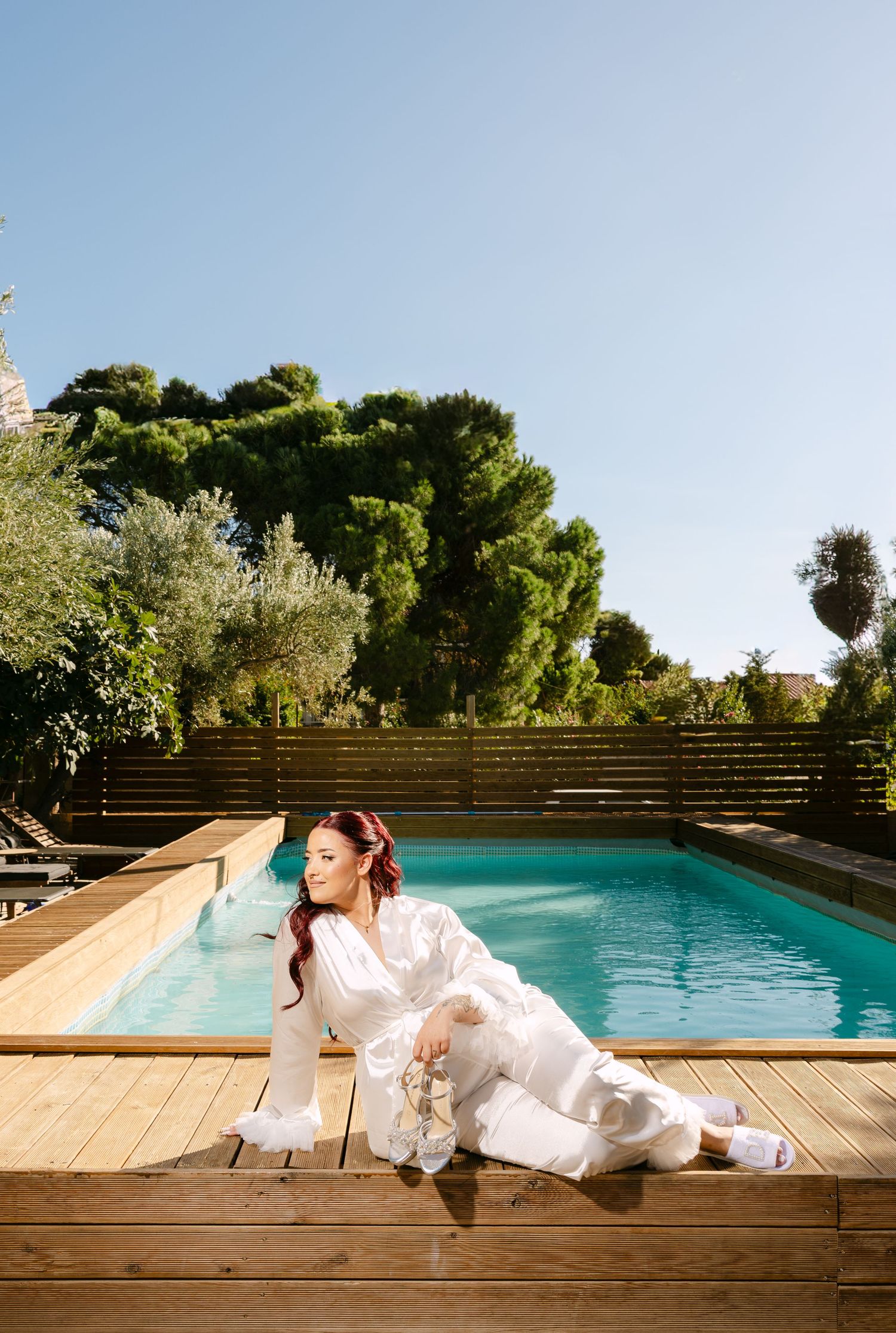 A bride in a flowing white dress lounges elegantly by a modern swimming pool on a sunny afternoon.