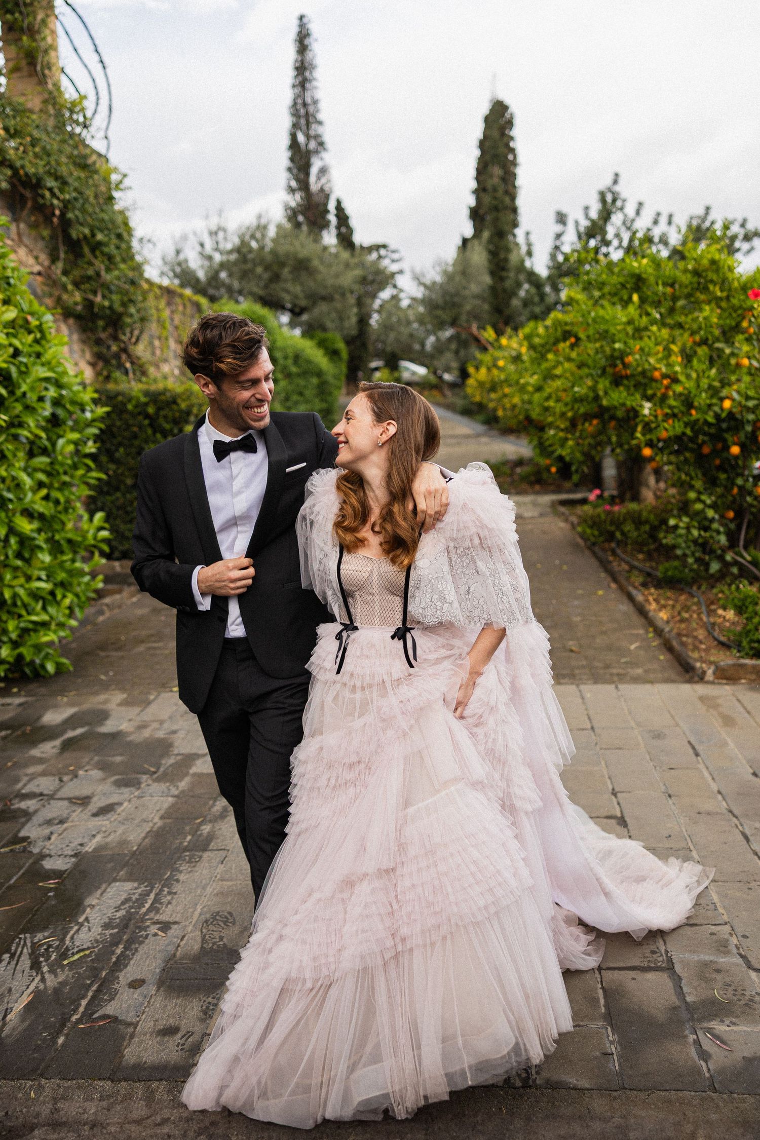A couple in wedding attire walk together along a garden path lined with lush greenery and tall cypress trees.