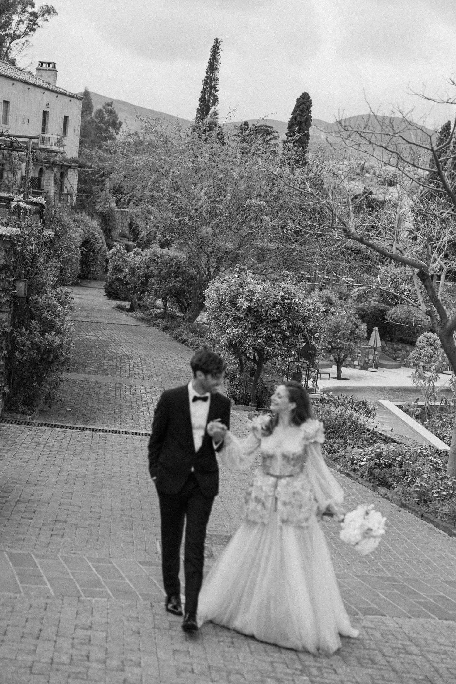 A wedding couple walks together on a brick pathway surrounded by lush gardens and trees at a historic venue.