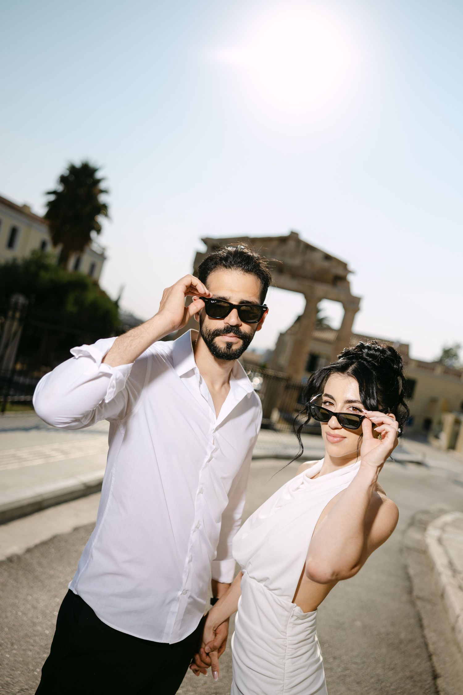 A couple in white outfits and sunglasses pose playfully in a series of photos against a historic European backdrop.