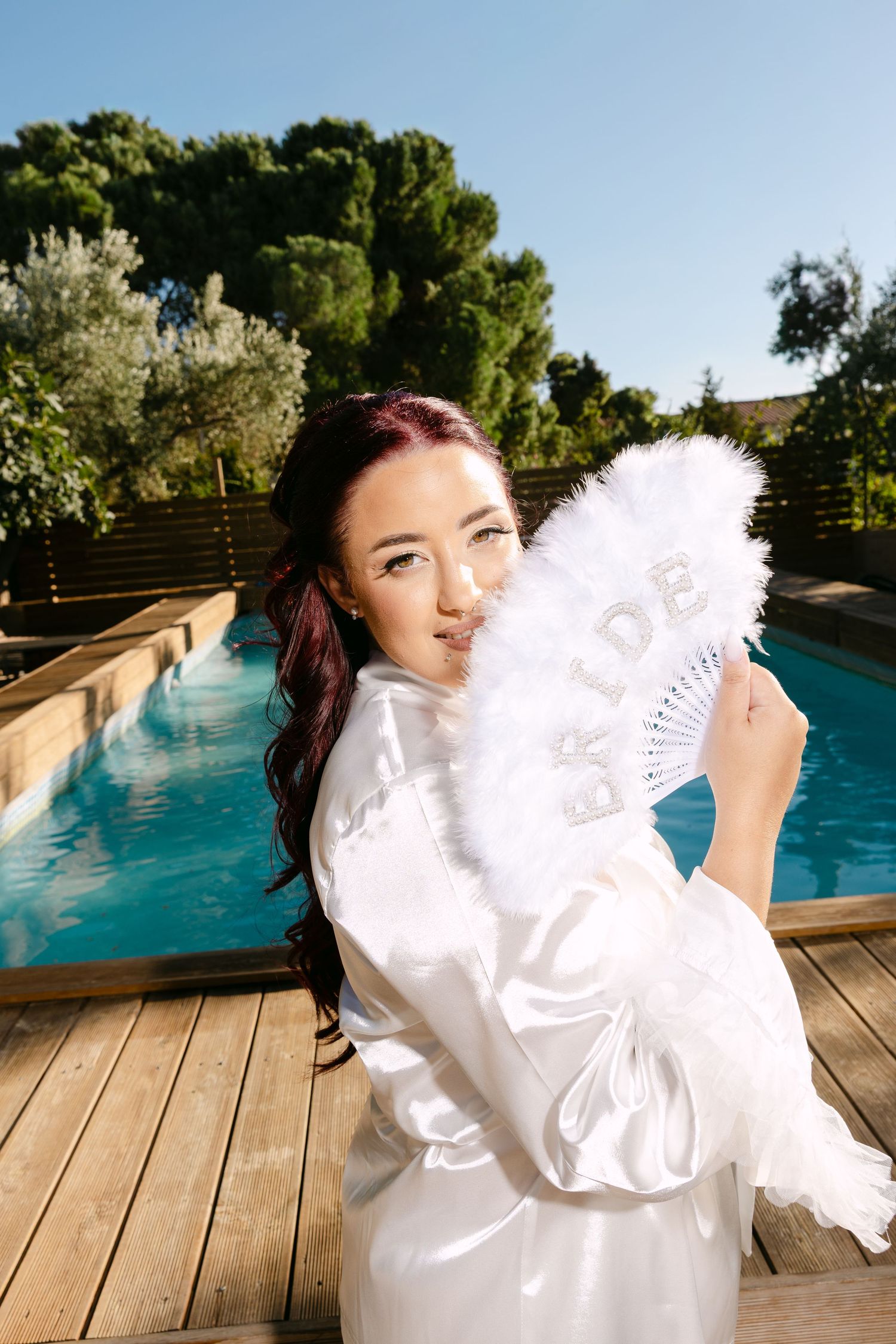 A person in white holds a decorative lace fan while posing by a swimming pool on a wooden deck.