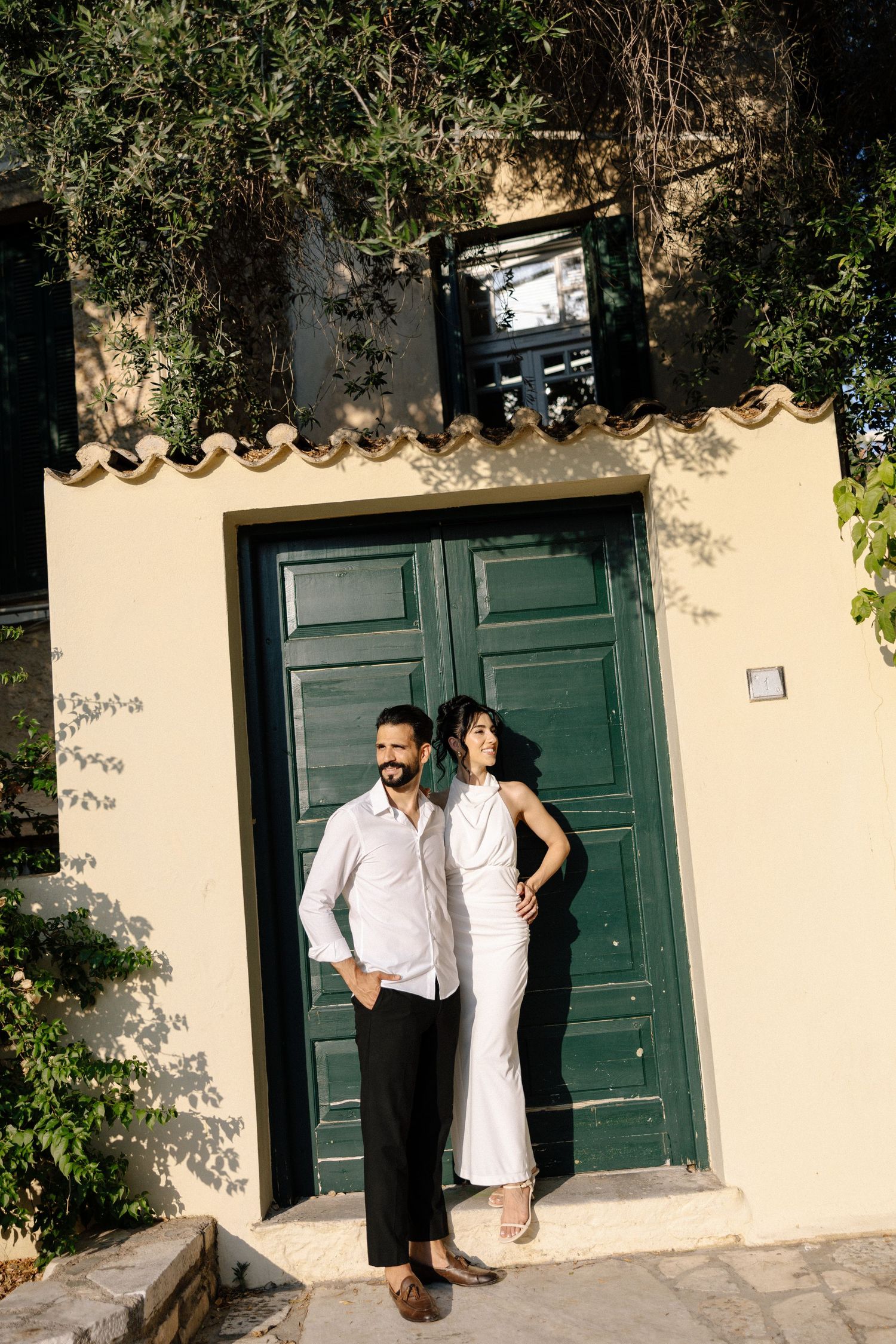 A couple poses in front of a large green door on a Mediterranean style building with climbing vines.