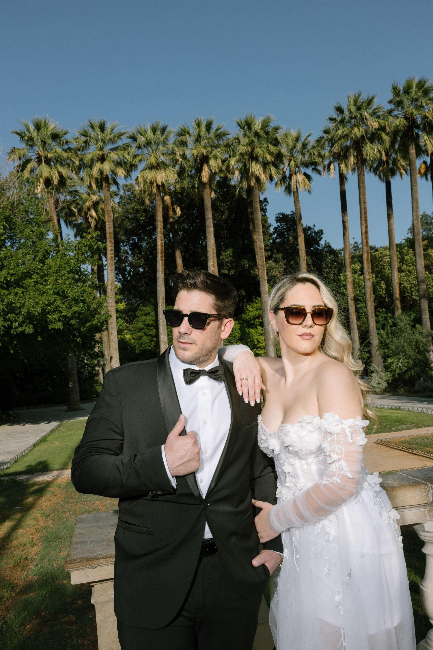 Wedding couple poses together in front of tall palm trees at an outdoor venue on a sunny day.