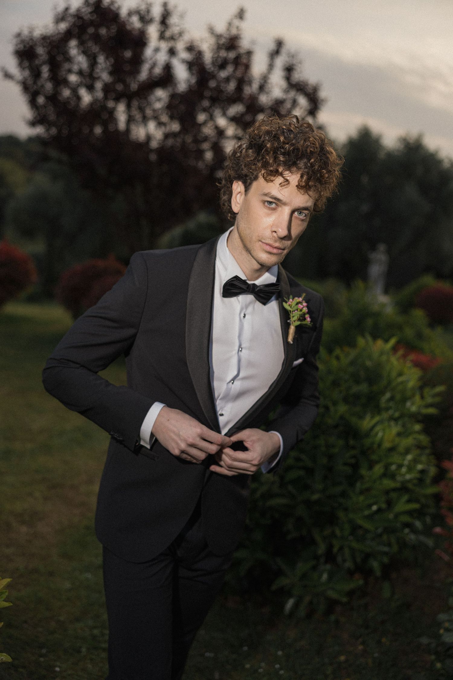 A person in a formal black tuxedo and bowtie poses in an outdoor garden setting at dusk.