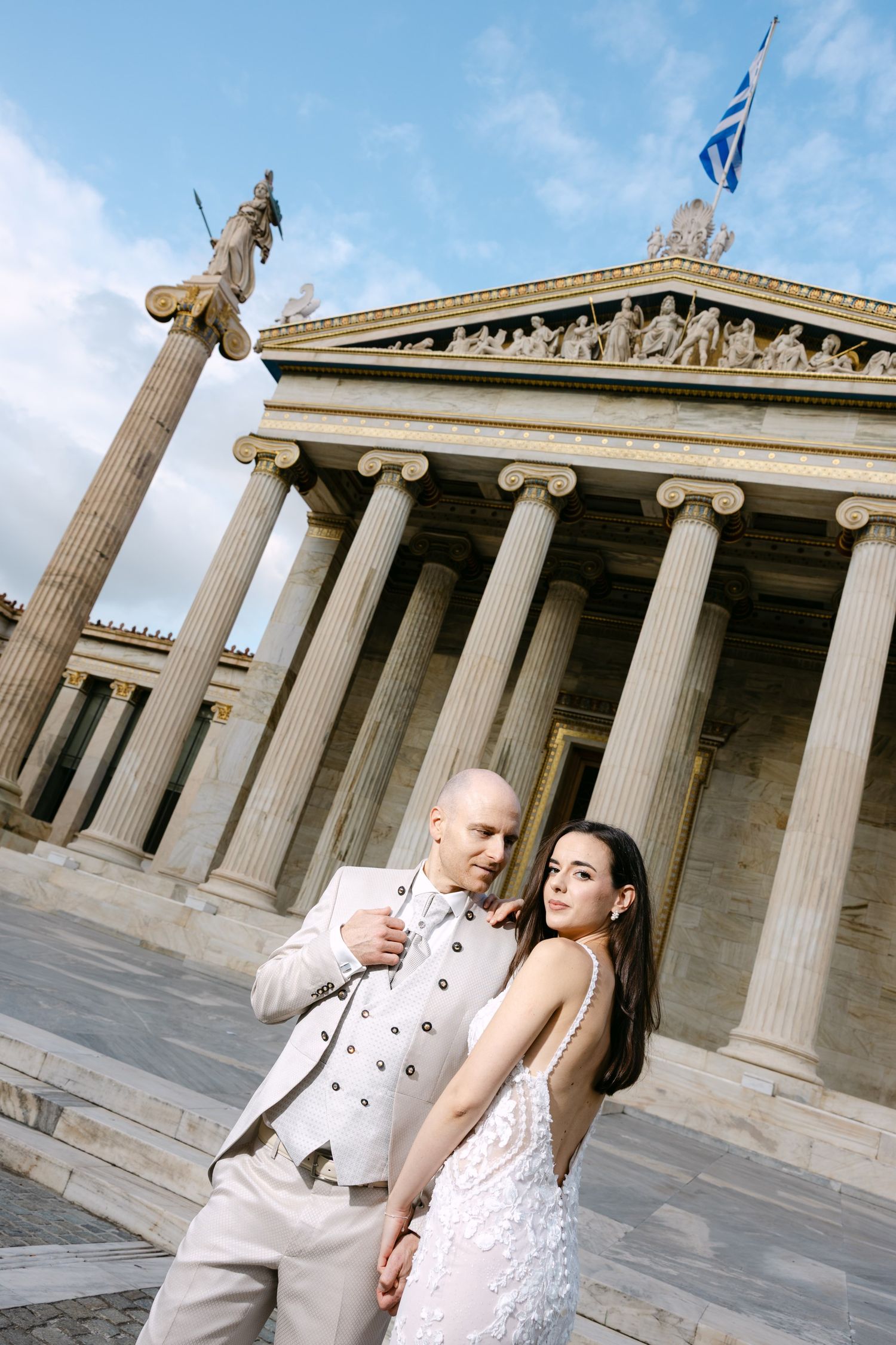 Elegant couple in formal attire pose for romantic wedding photos in front of a grand neoclassical building with columns.