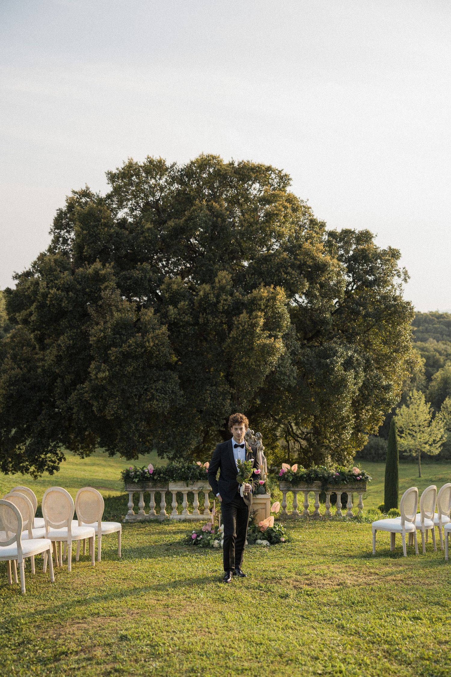 Outdoor ceremony setup on a grassy hillside with white chairs and mature oak trees in the background.
