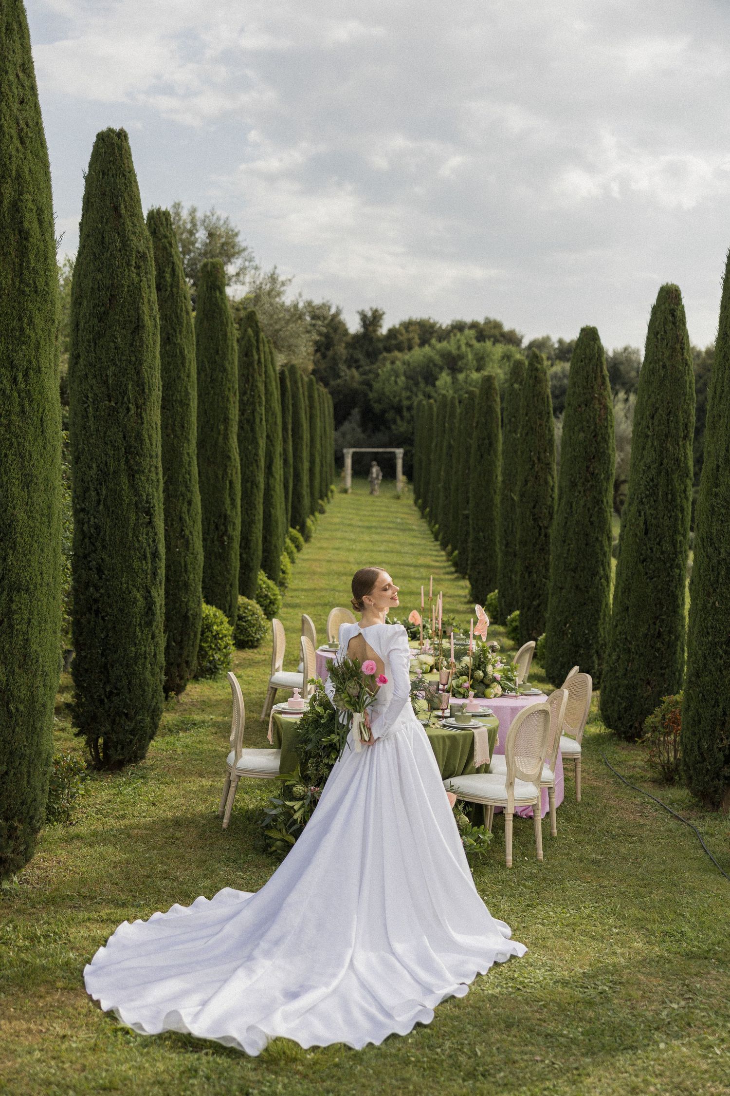 An elegant outdoor wedding table setting nestled between rows of tall cypress trees in a formal garden.